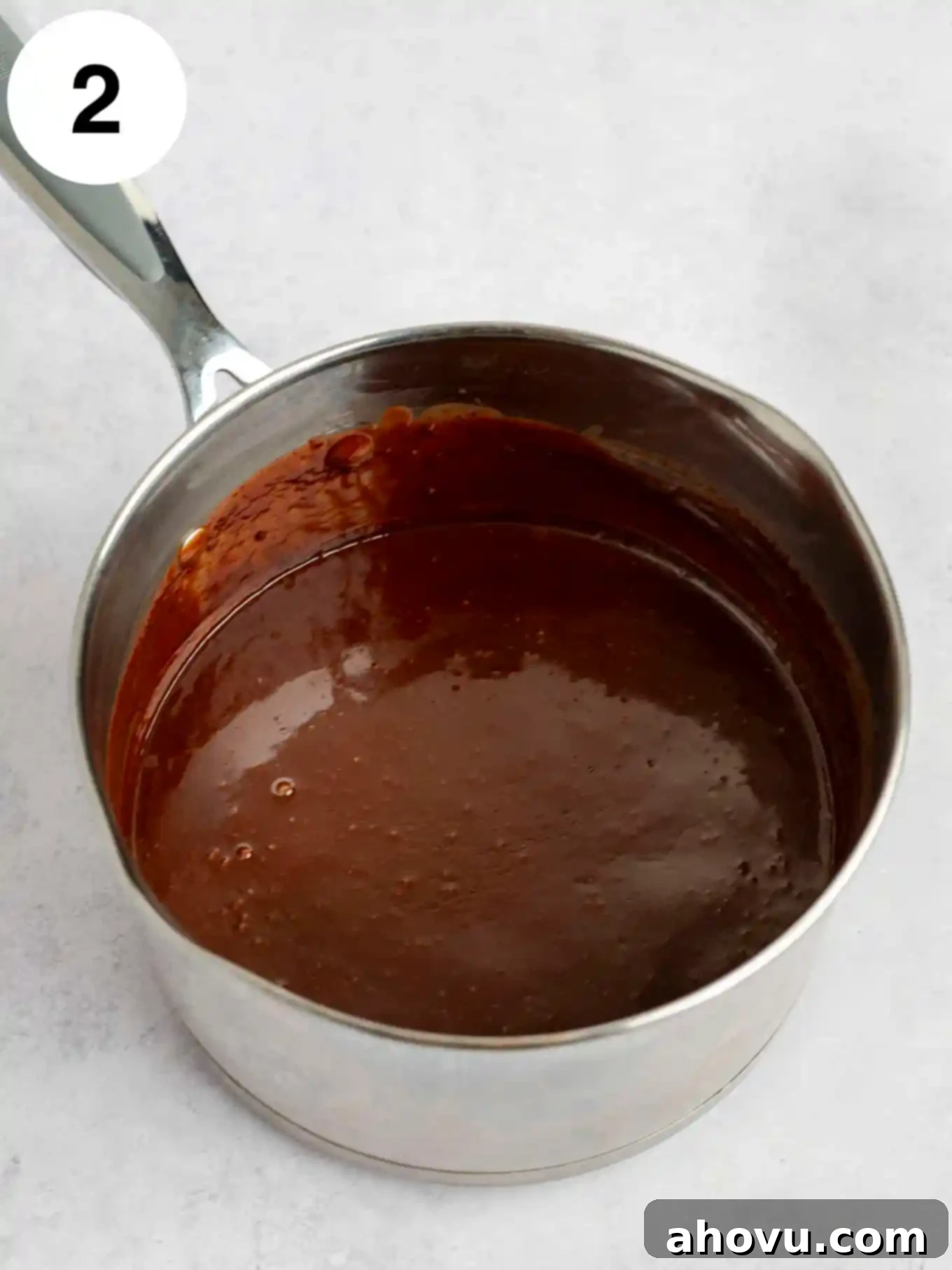 A bubbling metal saucepan on the stovetop, containing a smooth, dark, and glossy classic hot fudge sauce, just finished simmering.
