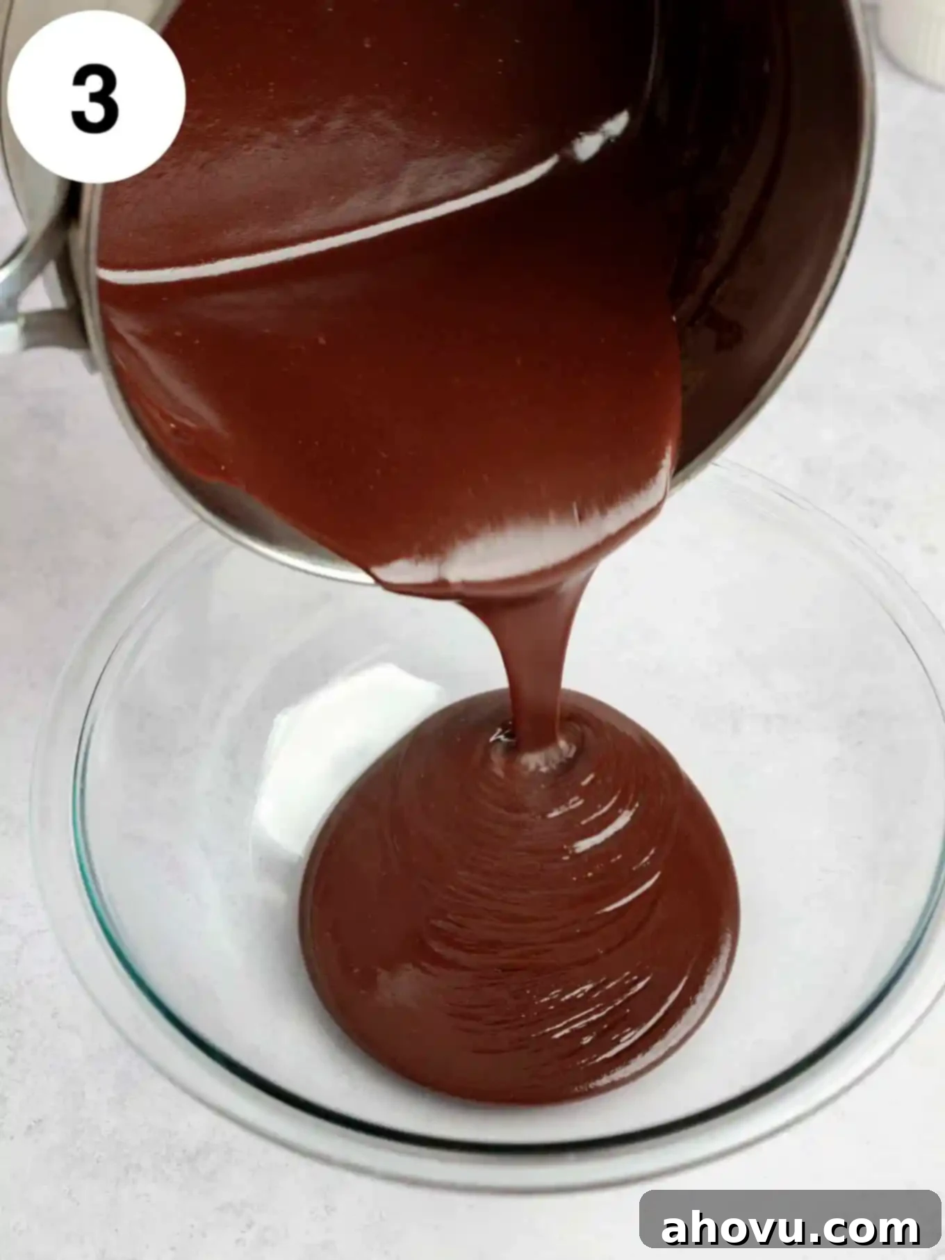 An action shot capturing a thick stream of rich, homemade hot fudge sauce being poured from a saucepan into a clear glass bowl, showcasing its smooth texture.
