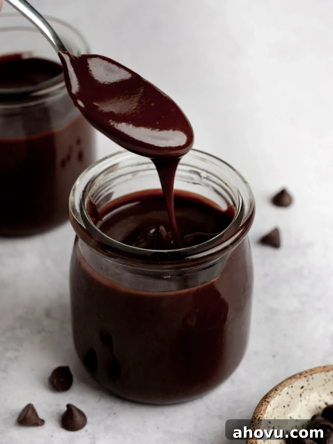A metal spoon lifting a generous dollop of glossy hot fudge sauce, suspended over a glass jar filled with more of the delicious homemade topping.