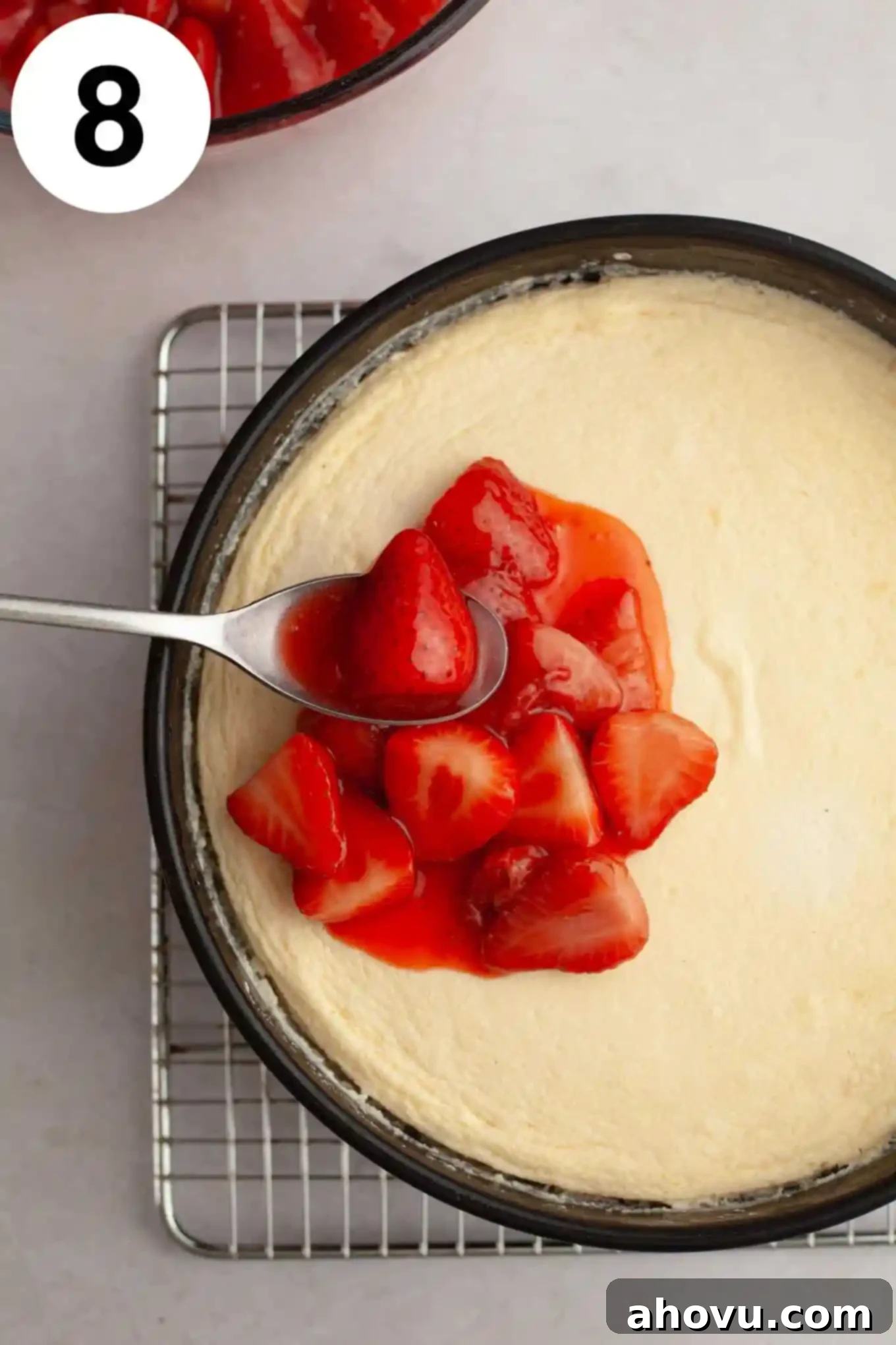 An overhead shot depicting thick, vibrant strawberry topping being generously spooned over a perfectly baked and cooled cheesecake.
