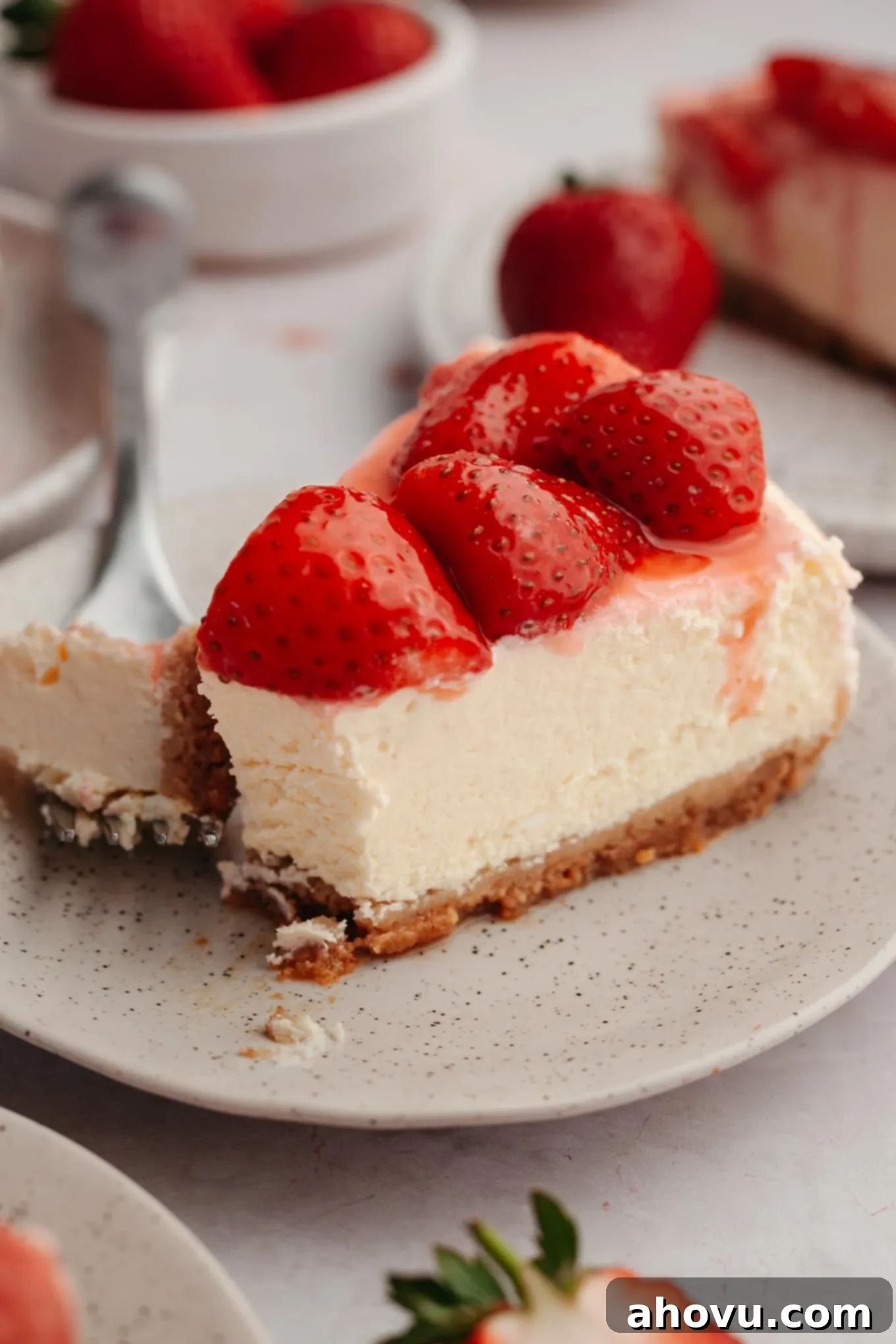 A close-up view of a strawberry cheesecake slice on a white plate, with a fork gently piercing a bite of the creamy dessert.