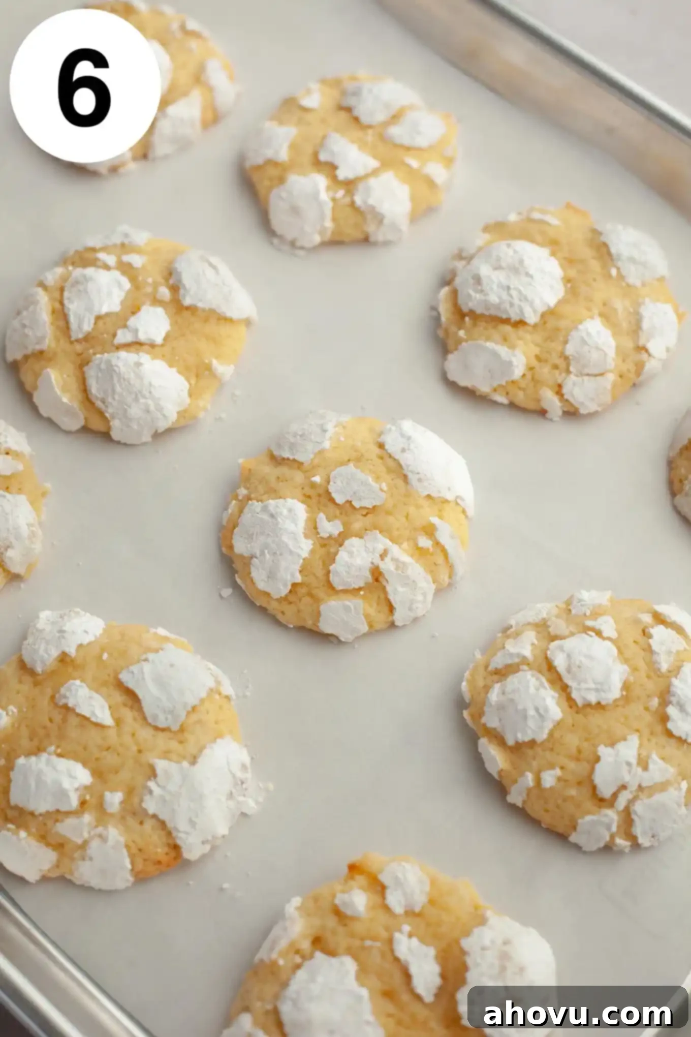 A fresh batch of baked lemon crinkle cookies displaying beautiful, defined cracks and a bright white powdered sugar coating on a parchment paper-lined baking sheet.