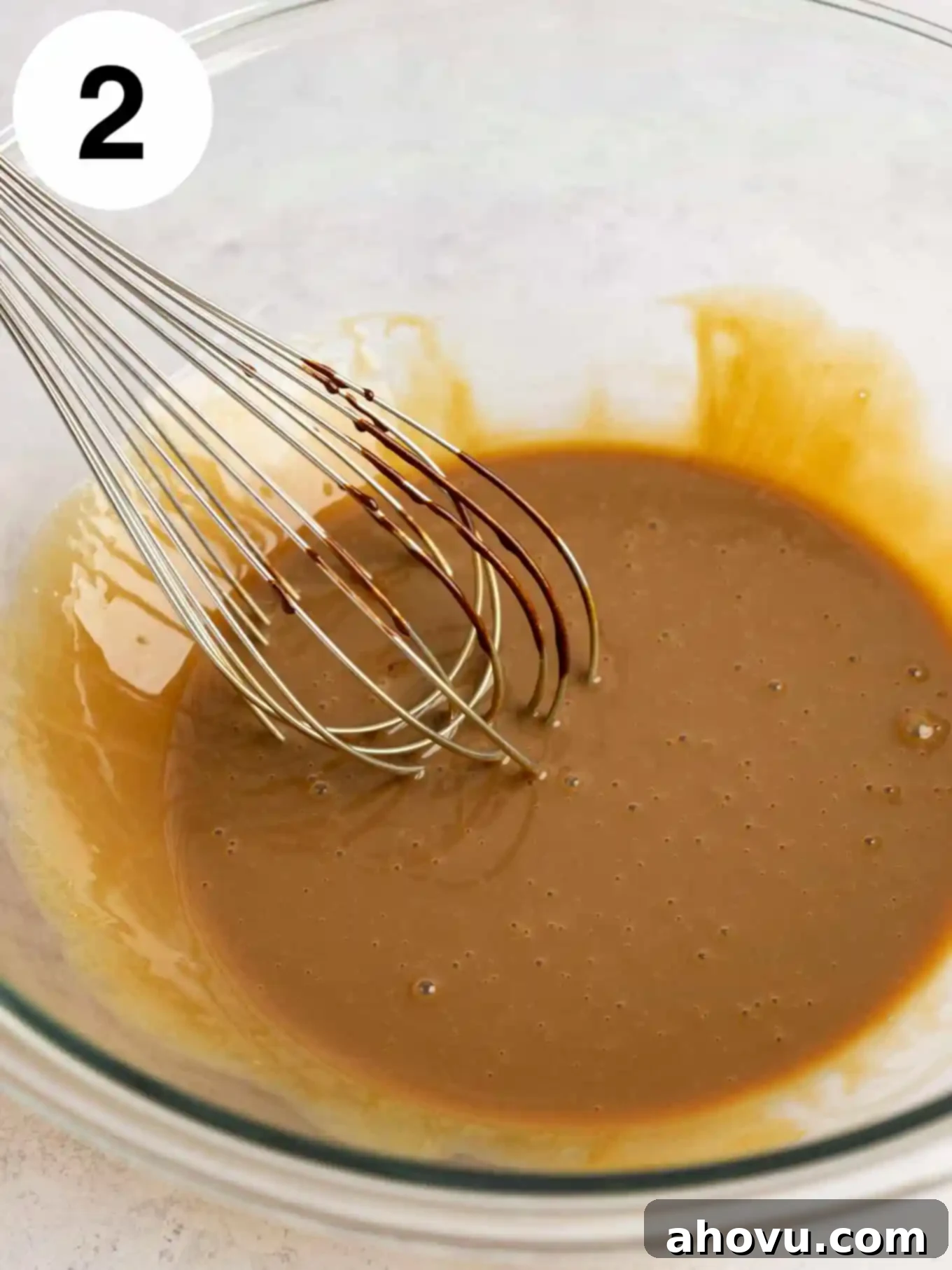 A glass mixing bowl with the coffee-condensed milk base mixture, ready for the whipped cream to be folded in, with a whisk resting in the bowl.