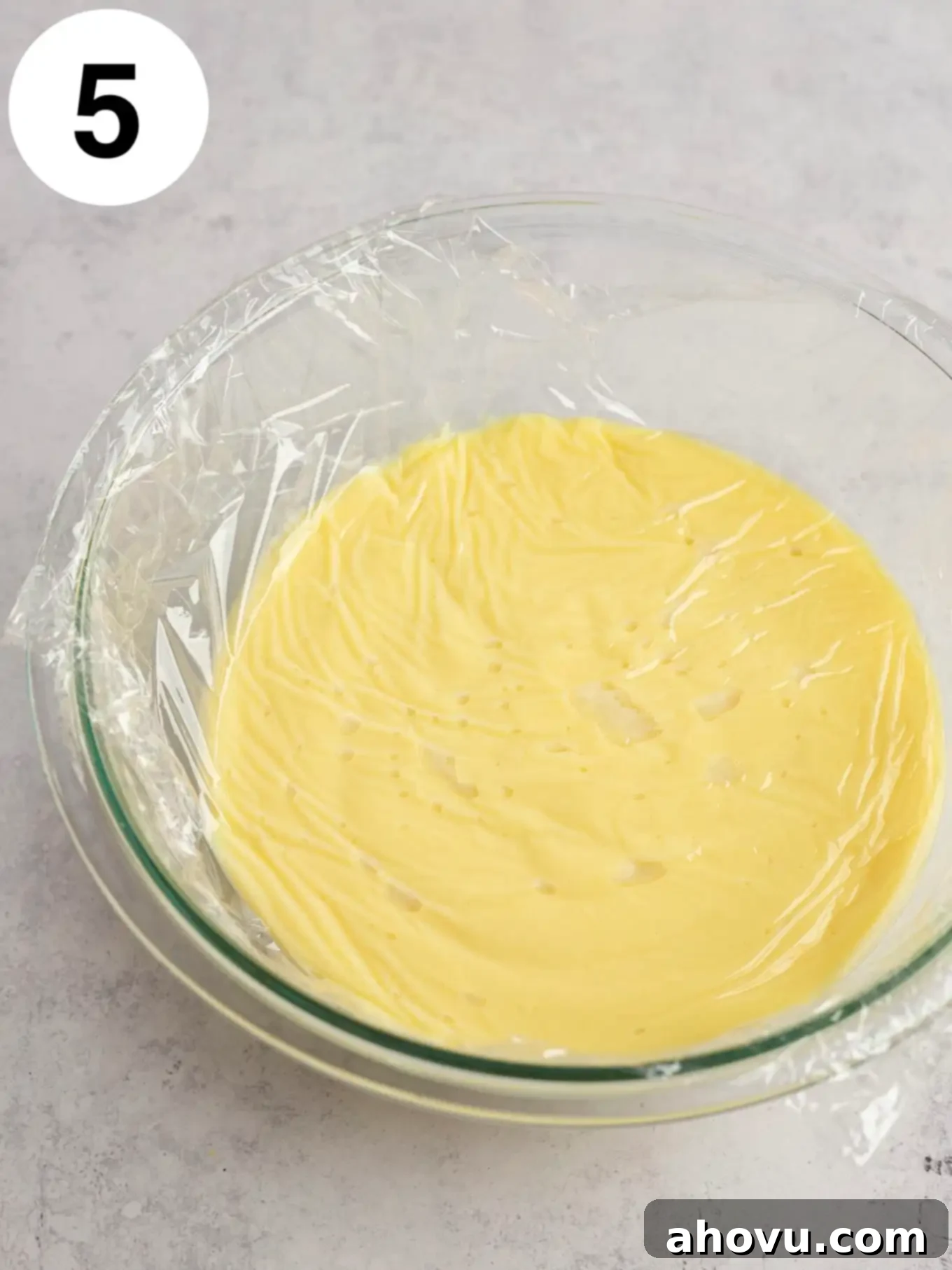 Creamy Vanilla Dream 8 Vanilla pudding in a glass bowl, with a piece of plastic wrap pressed directly on top of its surface to prevent a skin from forming during cooling, showcasing proper storage technique.