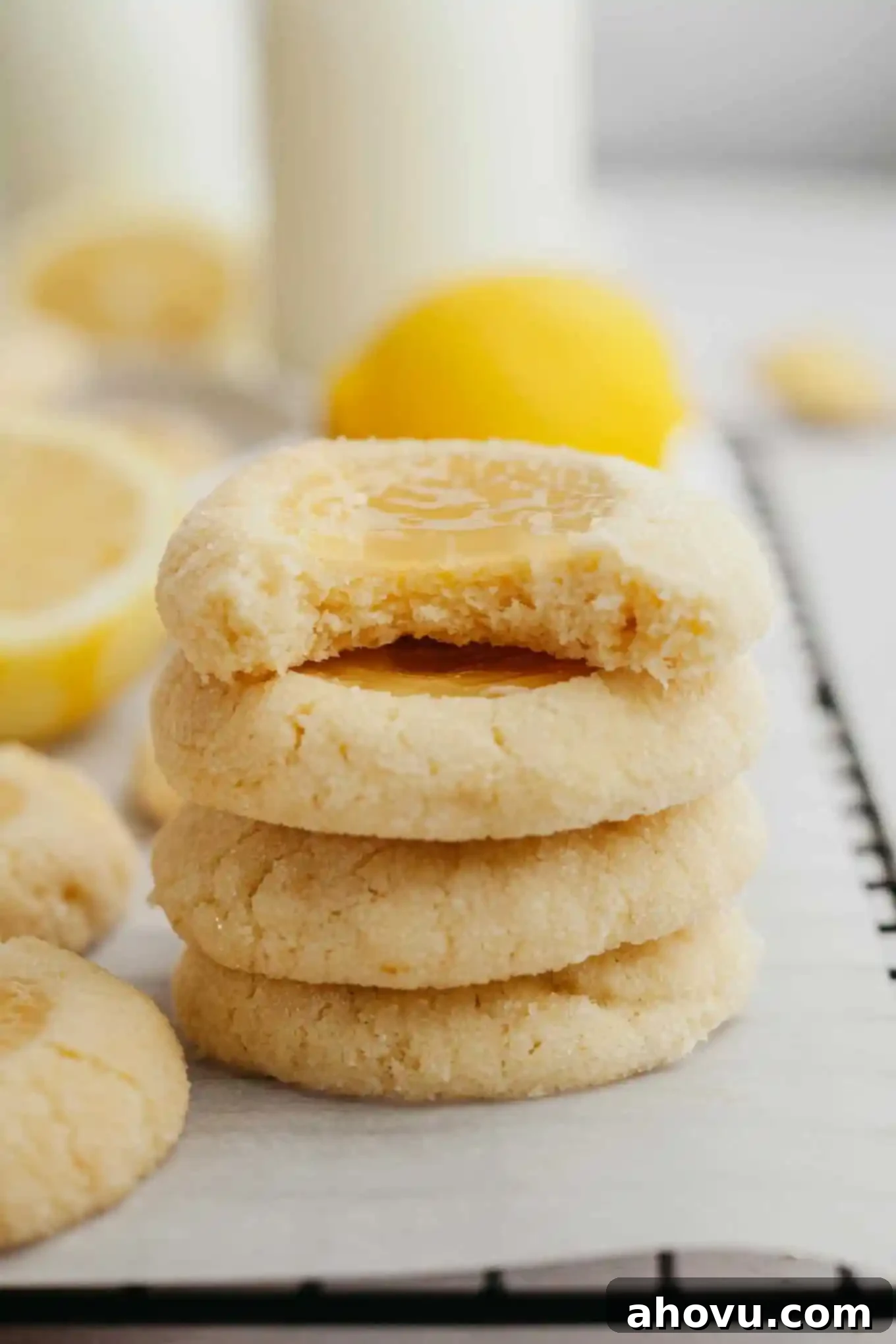A stack of four perfectly baked lemon curd thumbprint cookies. The top cookie has a bite taken out, revealing the vibrant yellow lemon curd filling.