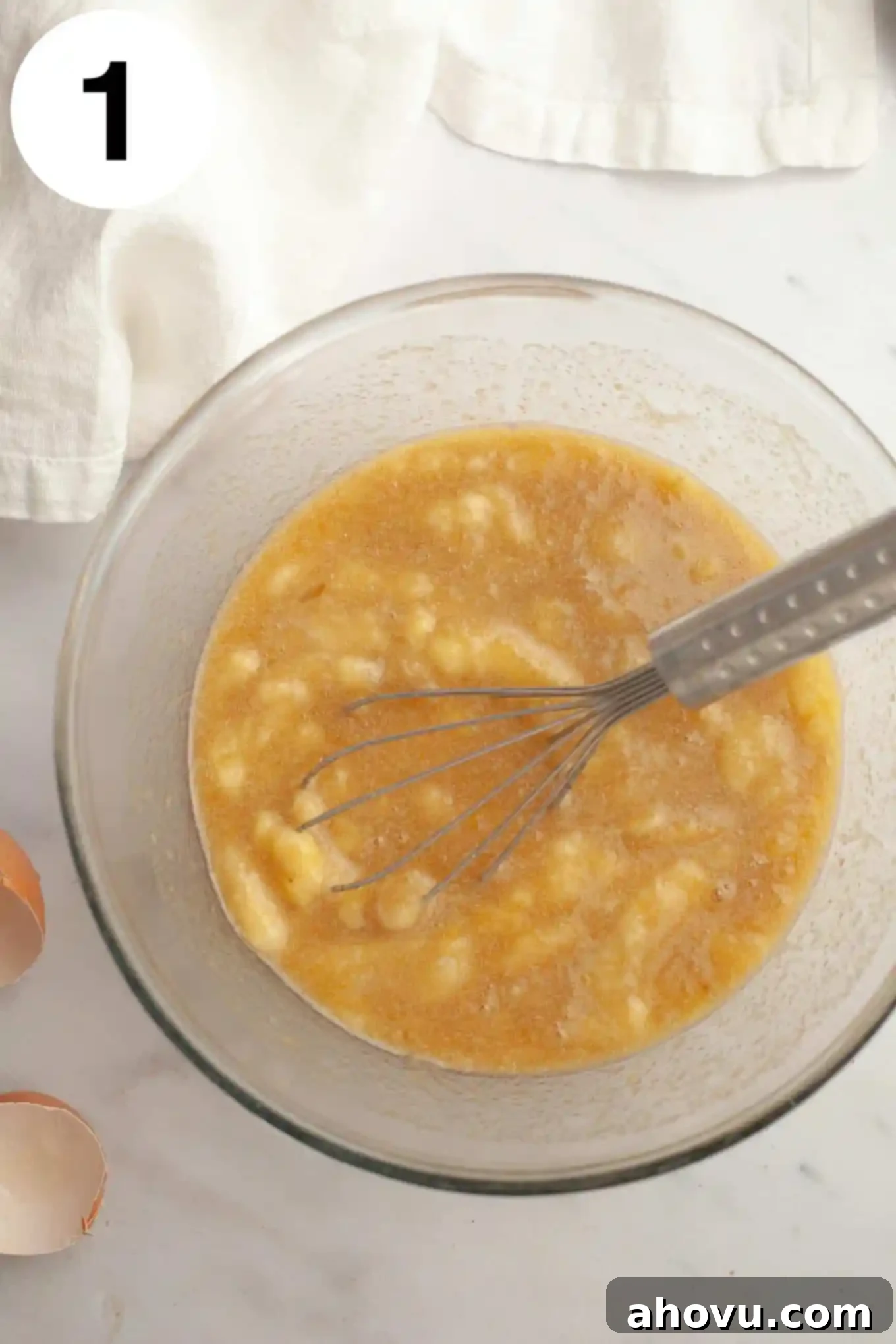 An overhead view of a banana and egg mixture in a glass mixing bowl, being whisked to combine the wet ingredients for the muffins.