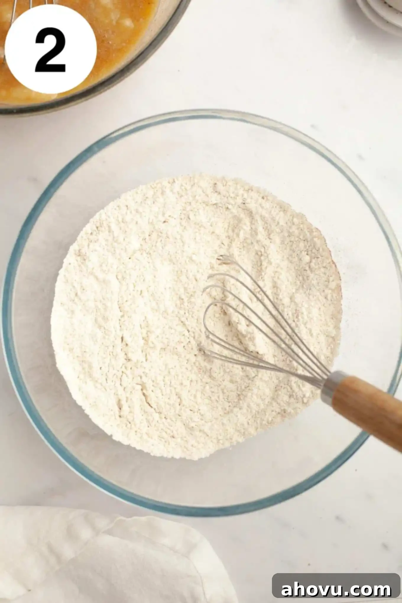 An overhead view of a flour mixture in a glass mixing bowl, with a whisk, showing the dry ingredients perfectly combined.