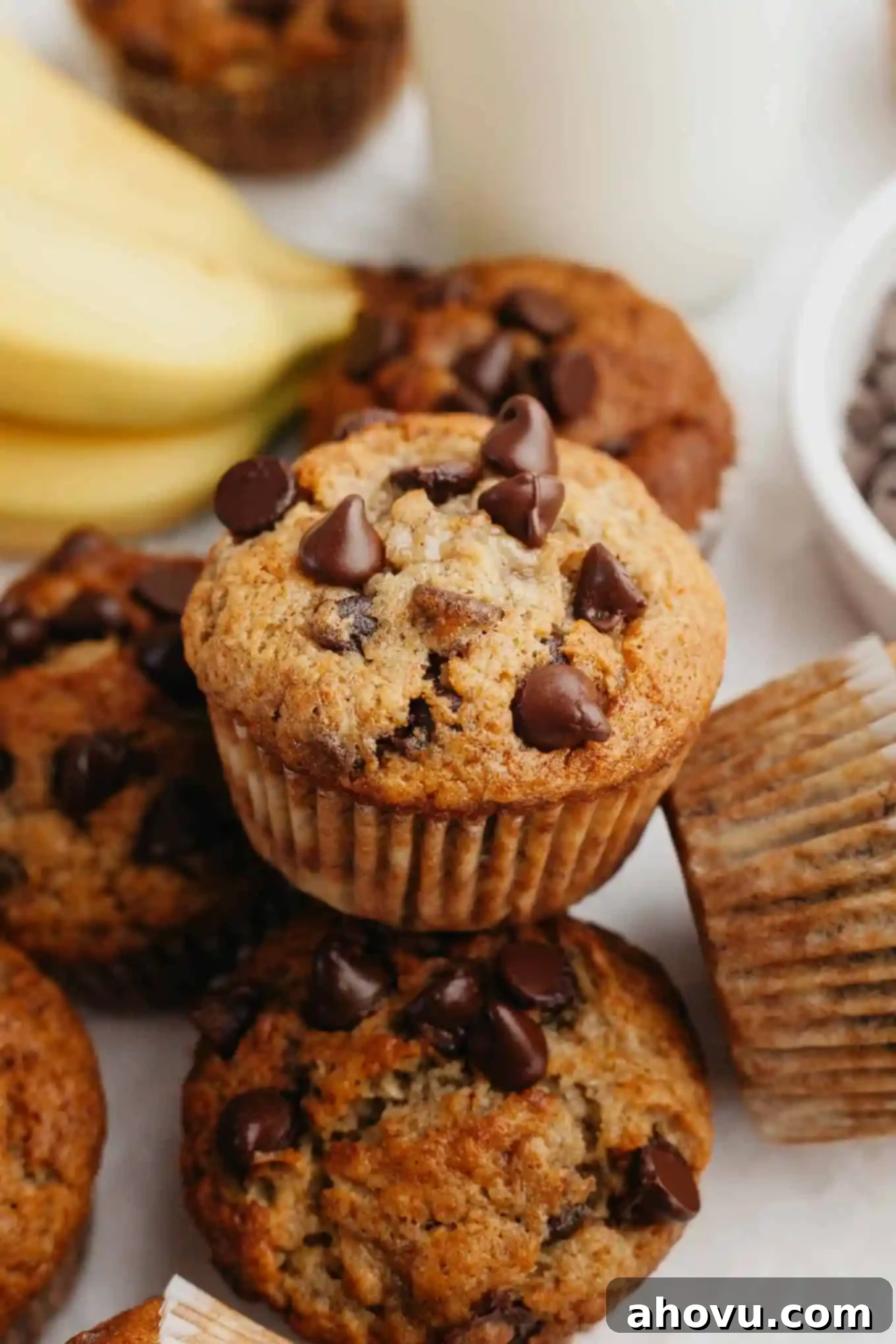 A close-up view of a perfectly baked banana and chocolate chip muffin, resting on a pile of other delicious muffins, highlighting its golden crust and moist interior.