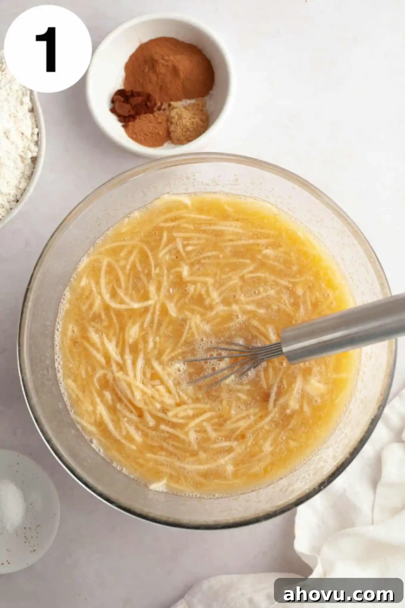 An overhead view of the wet ingredients, including grated apple, being whisked in a glass mixing bowl.