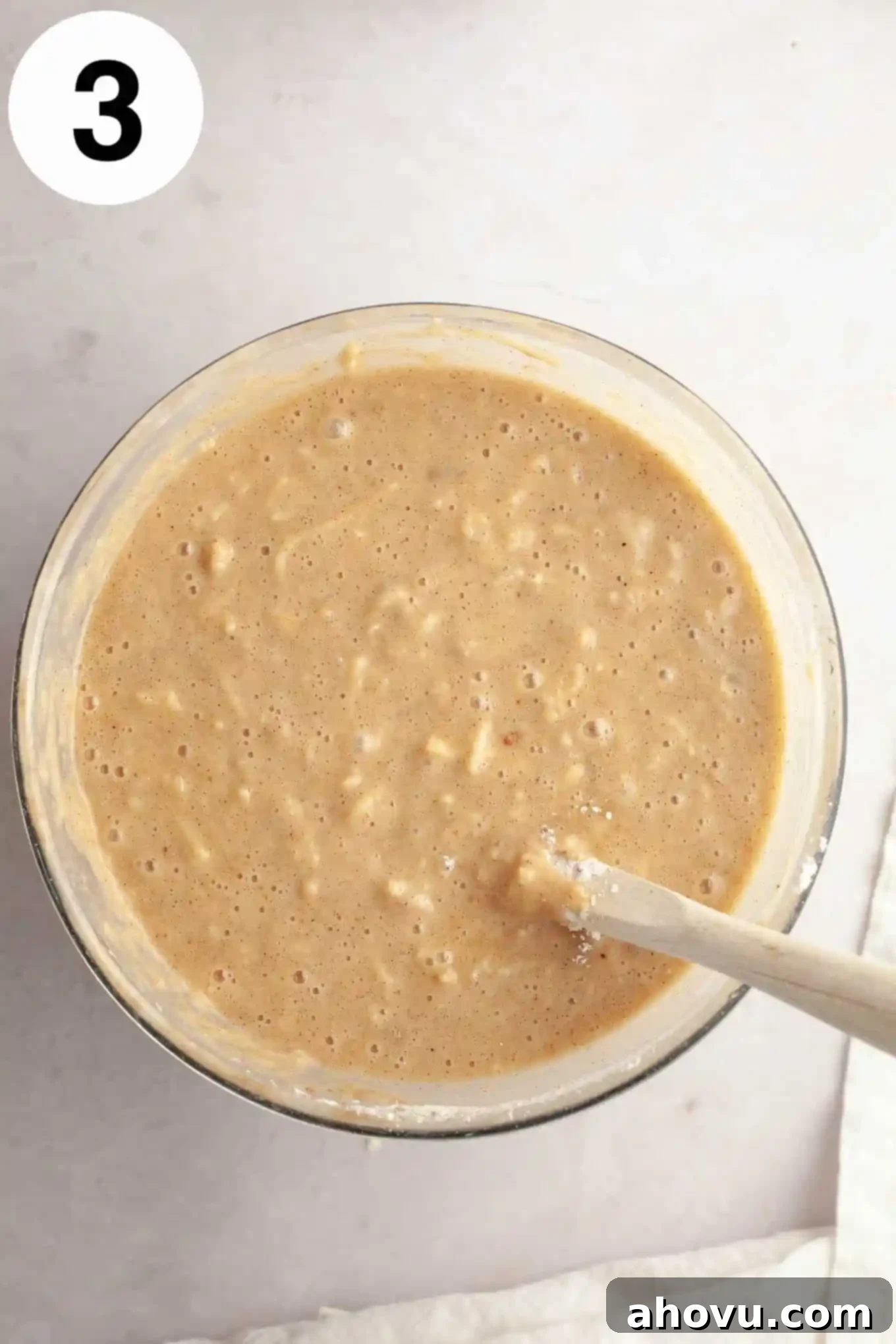 An overhead view of apple cake batter, perfectly mixed in a glass mixing bowl with a wooden spoon, ready for the bundt pan.