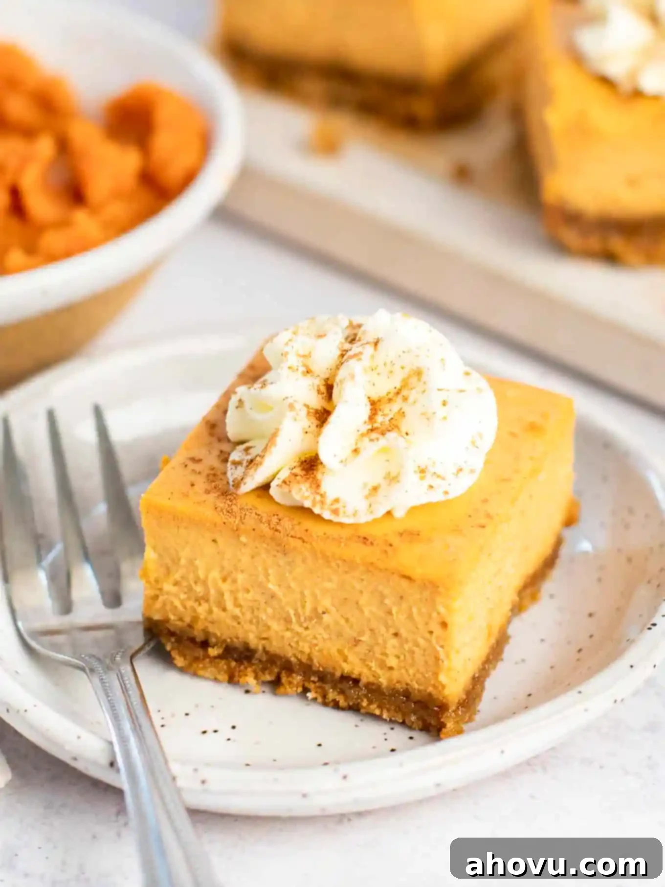 A close-up view of a single pumpkin cheesecake bar on a white dessert plate, beautifully garnished with a generous swirl of whipped cream and a sprinkle of pumpkin pie spice, inviting a delicious bite.