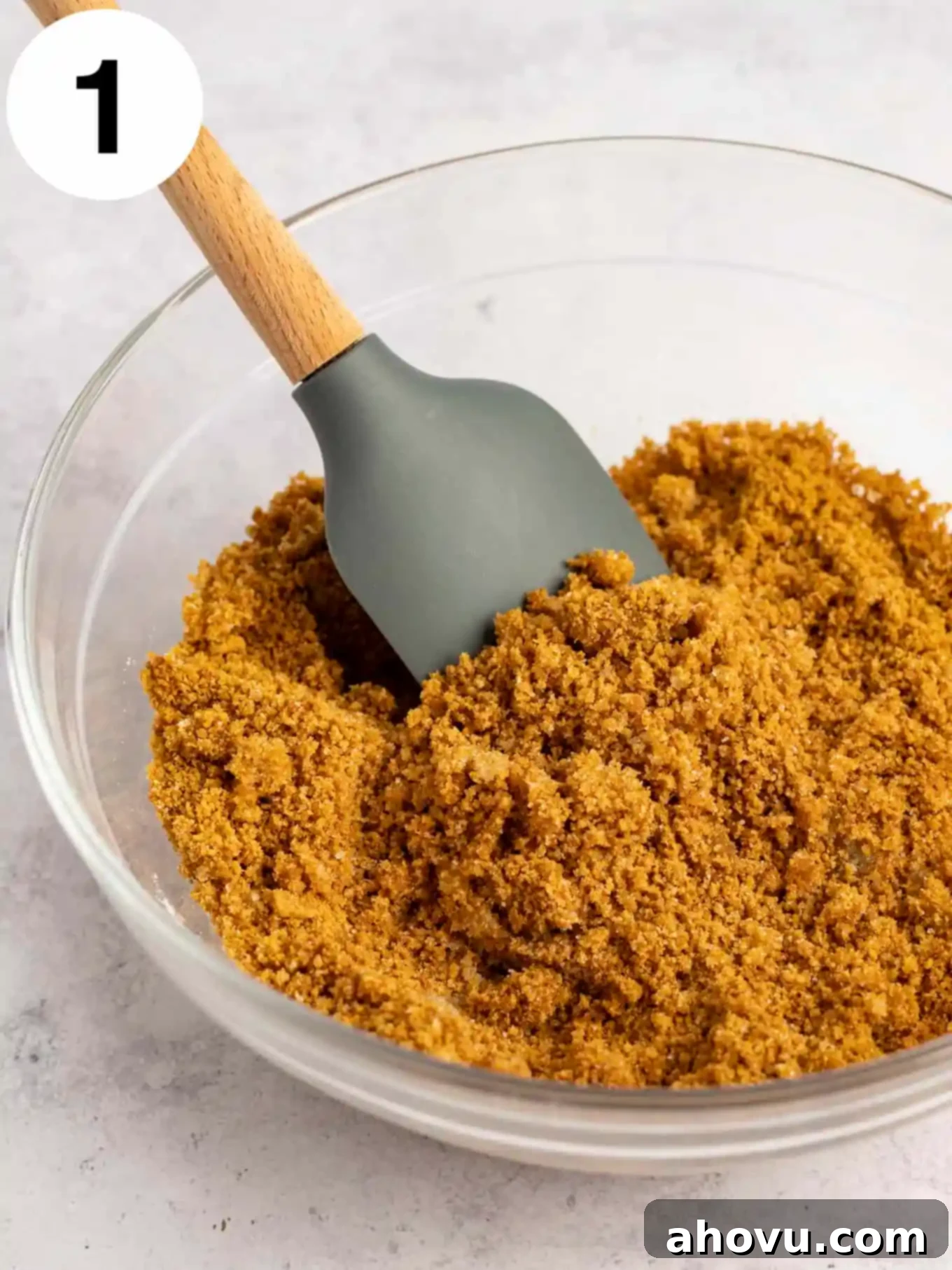 A glass mixing bowl holds a perfectly blended gingersnap cookie crumb mixture, prepared with melted butter and sugar, alongside a rubber spatula.