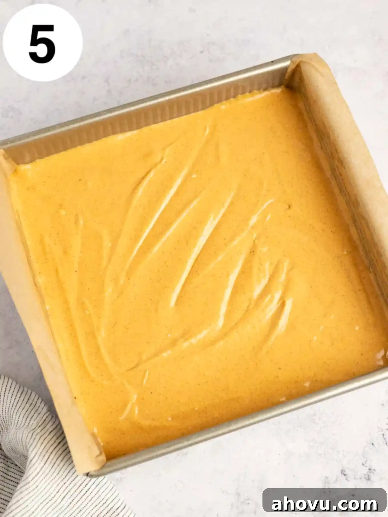 An overhead view of the unbaked pumpkin cheesecake filling perfectly spread over the gingersnap crust in a parchment-lined baking dish, ready for the oven.