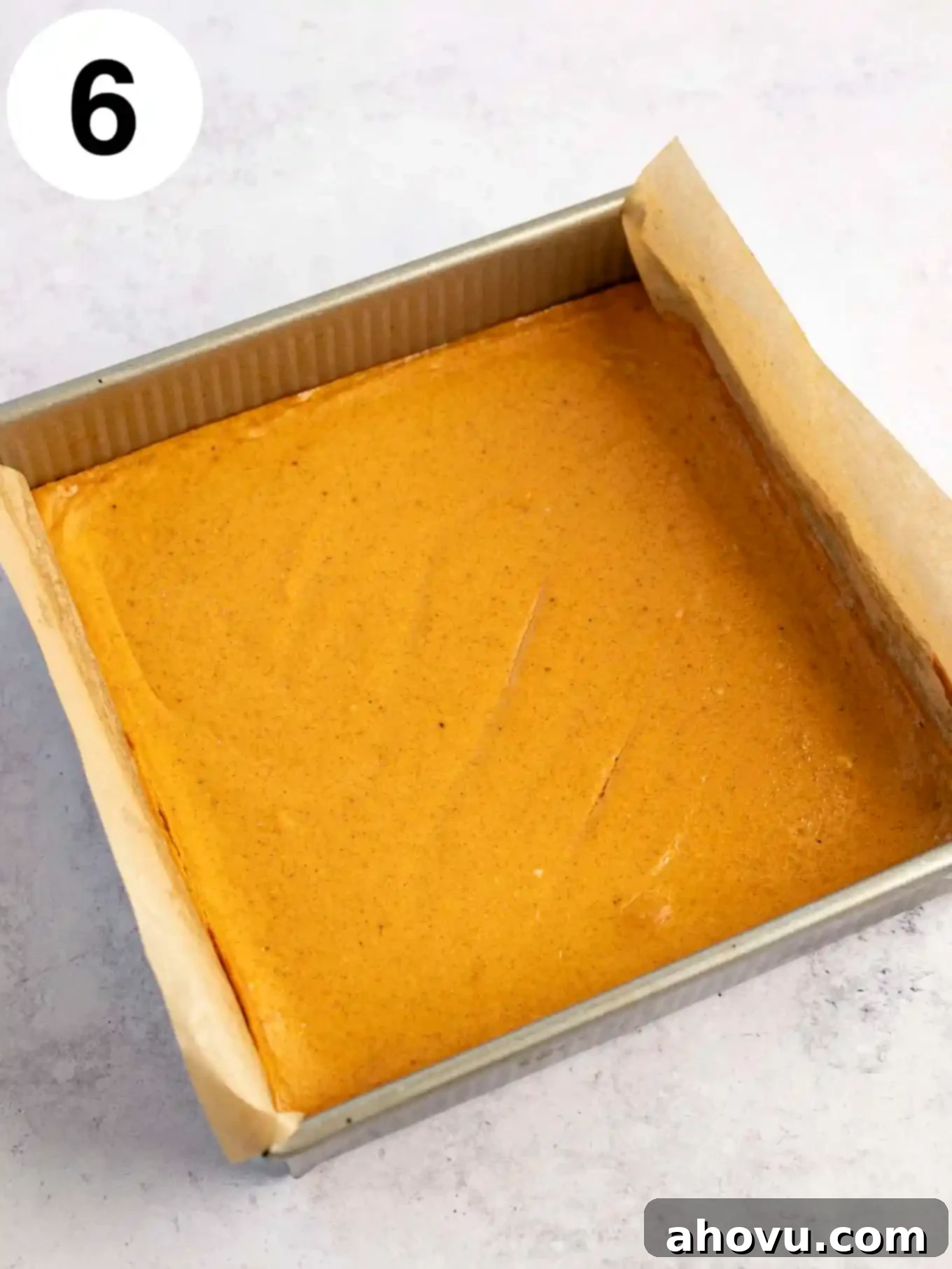 An overhead view of freshly baked pumpkin cheesecake bars cooling in a lined baking dish, their golden-brown edges and smooth surface hinting at their deliciousness.