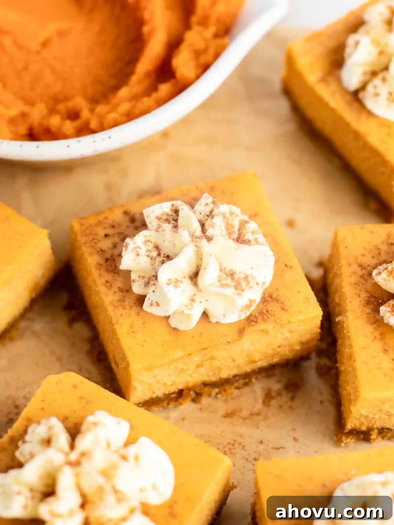 Several perfectly cut pumpkin cheesecake bars, elegantly arranged on parchment paper, adorned with swirls of whipped cream and a delicate dusting of pumpkin pie spice.