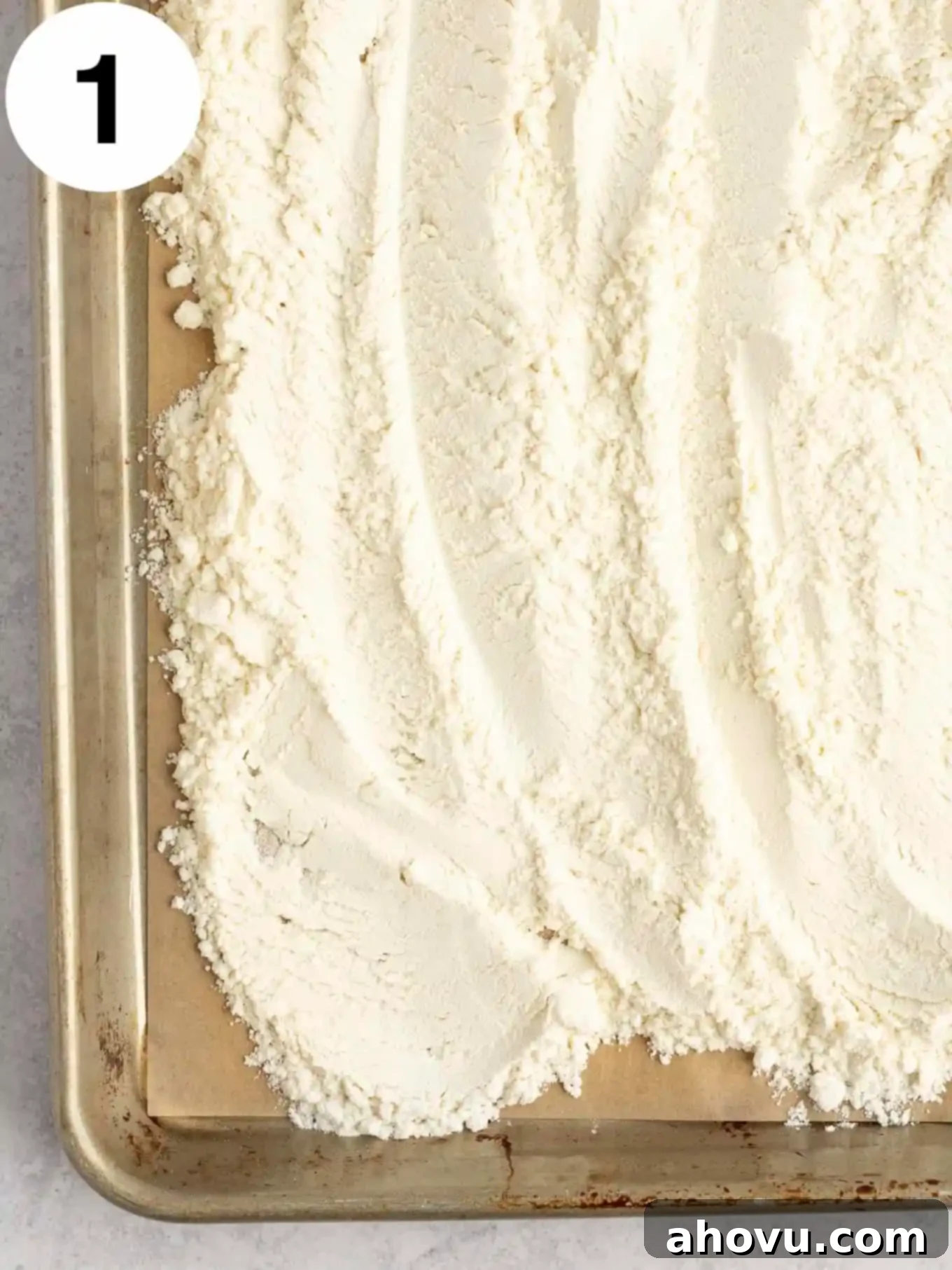 An overhead view of evenly spread heat-treated flour on a baking sheet, ready for cooling before being added to the frosting mixture.