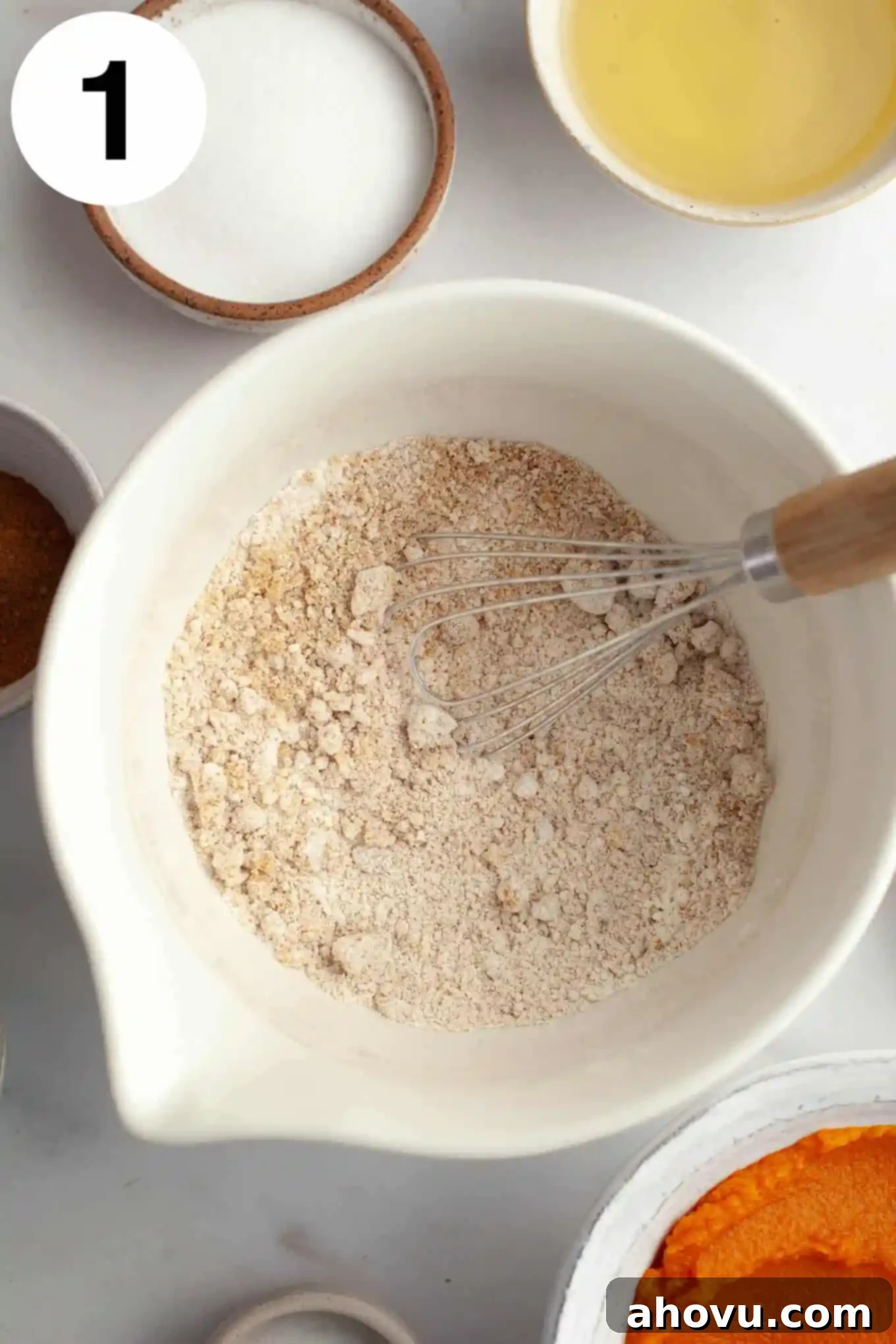 An overhead view of a crumbly flour mixture in a white mixing bowl, with a whisk. 