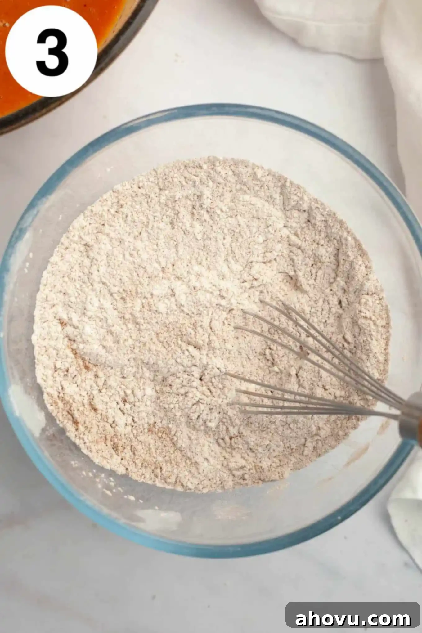 An overhead view of a dry flour mixture in a glass mixing bowl, with a whisk. 
