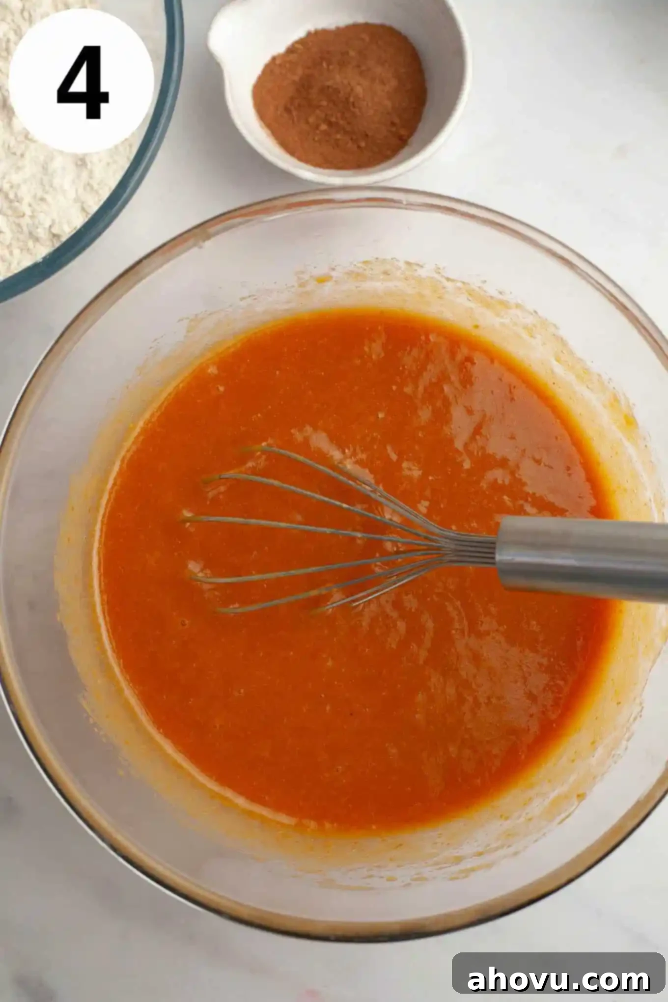 An overhead view of a wet pumpkin mixture in a glass mixing bowl, with a whisk. 