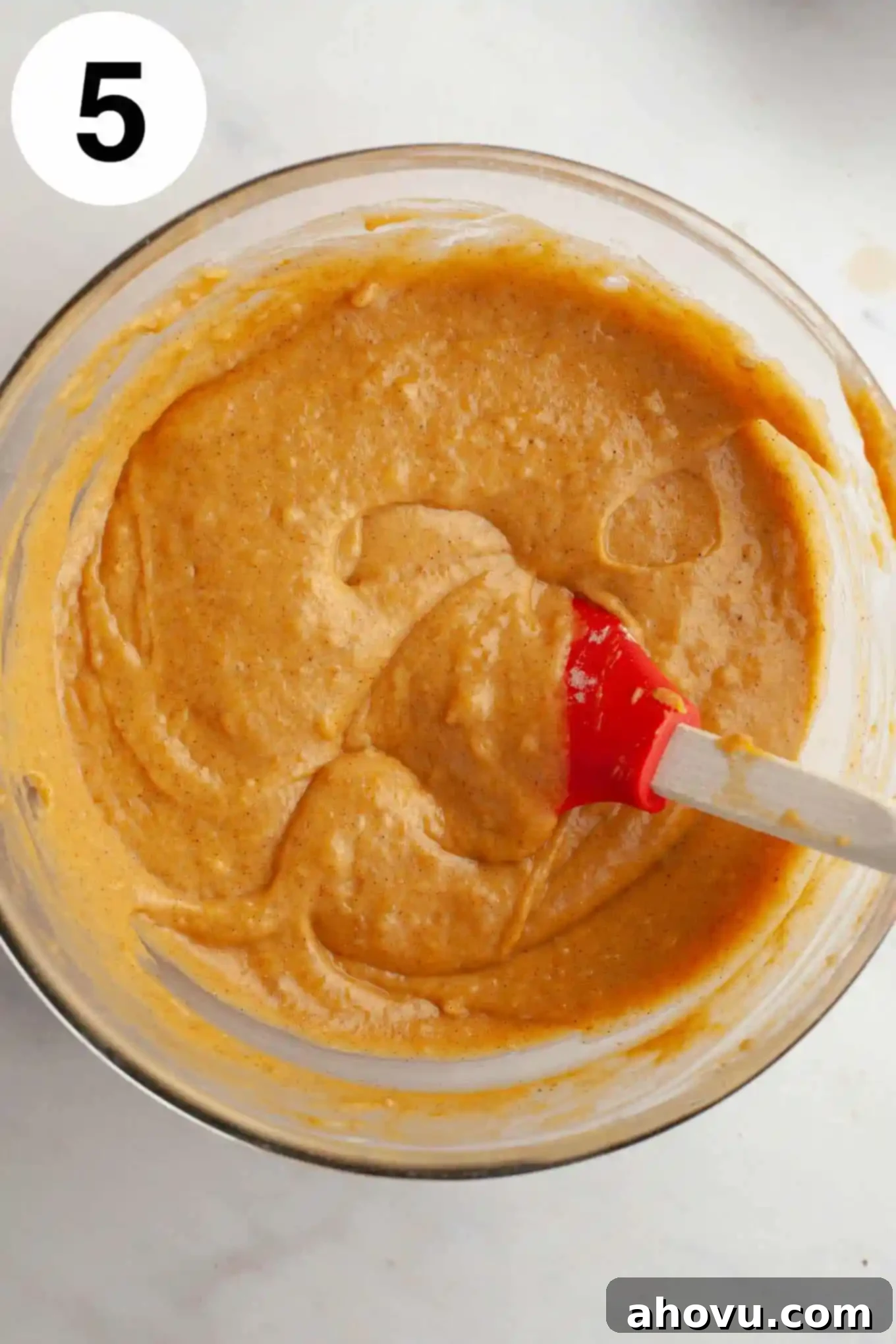 An overhead view of pumpkin cake batter in a glass mixing bowl, with a rubber spatula. 