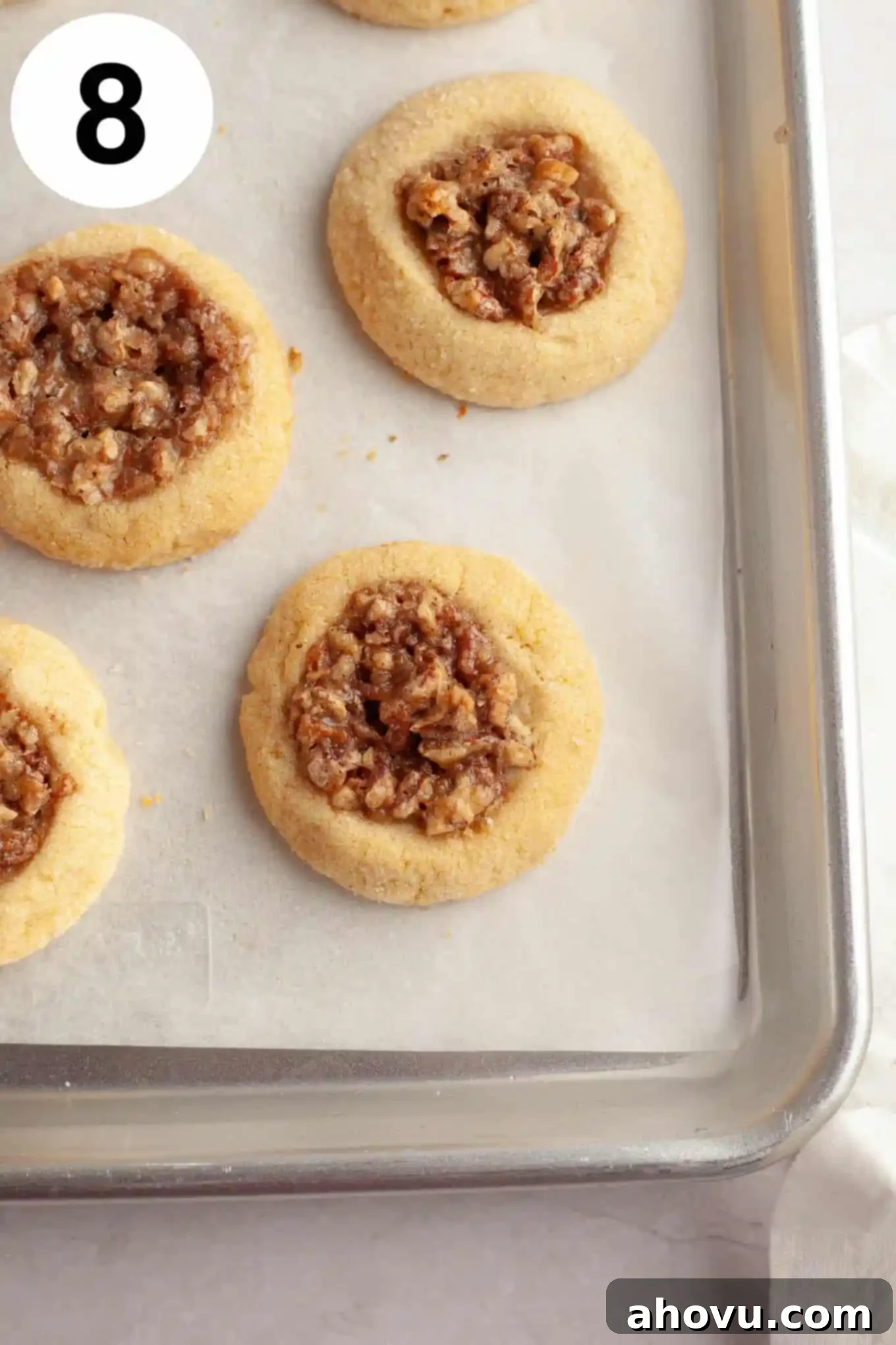 An overhead view of baked pecan pie cookies fresh out of the oven, cooling on a parchment paper-lined baking sheet, with the filling set and edges lightly browned. 