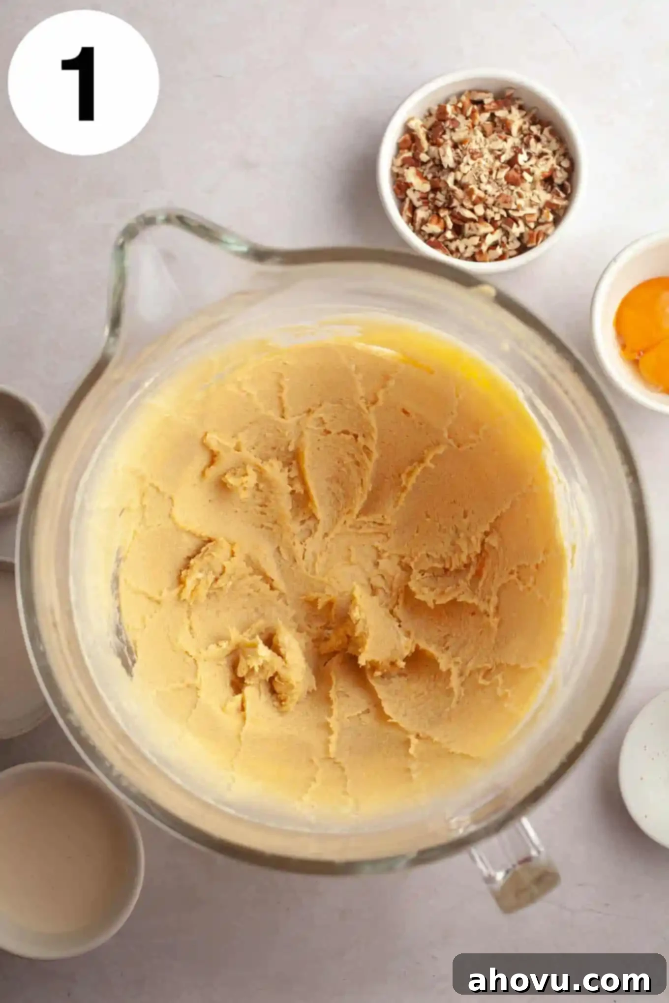 An overhead view of softened butter and light brown sugar being creamed together in a clear glass mixing bowl, forming a smooth, pale mixture. 