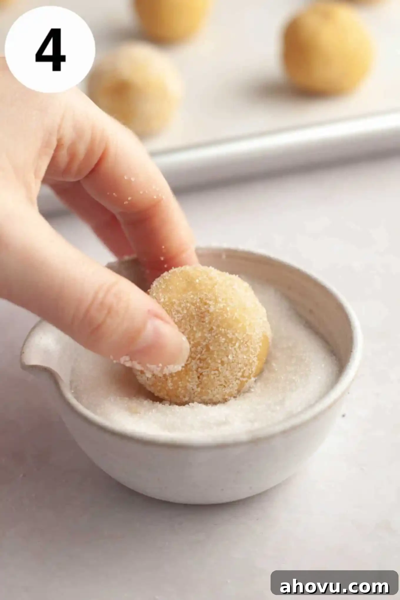 An action shot showing a portion of cookie dough being rolled between hands into a smooth ball, then coated in granulated sugar. 
