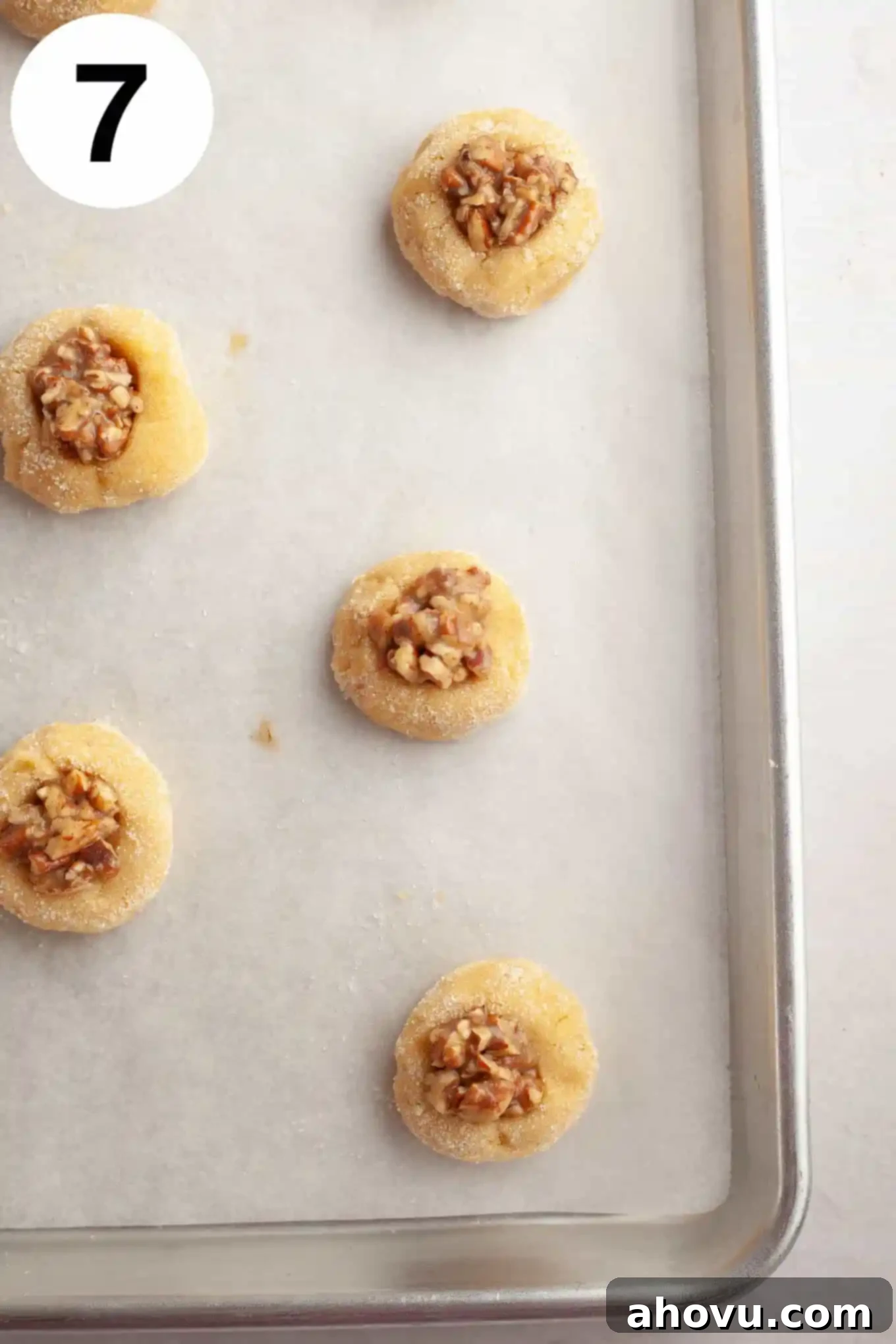 An overhead view of unbaked pecan pie cookies on a parchment paper-lined baking sheet, with each cookie filled with the pecan mixture. 