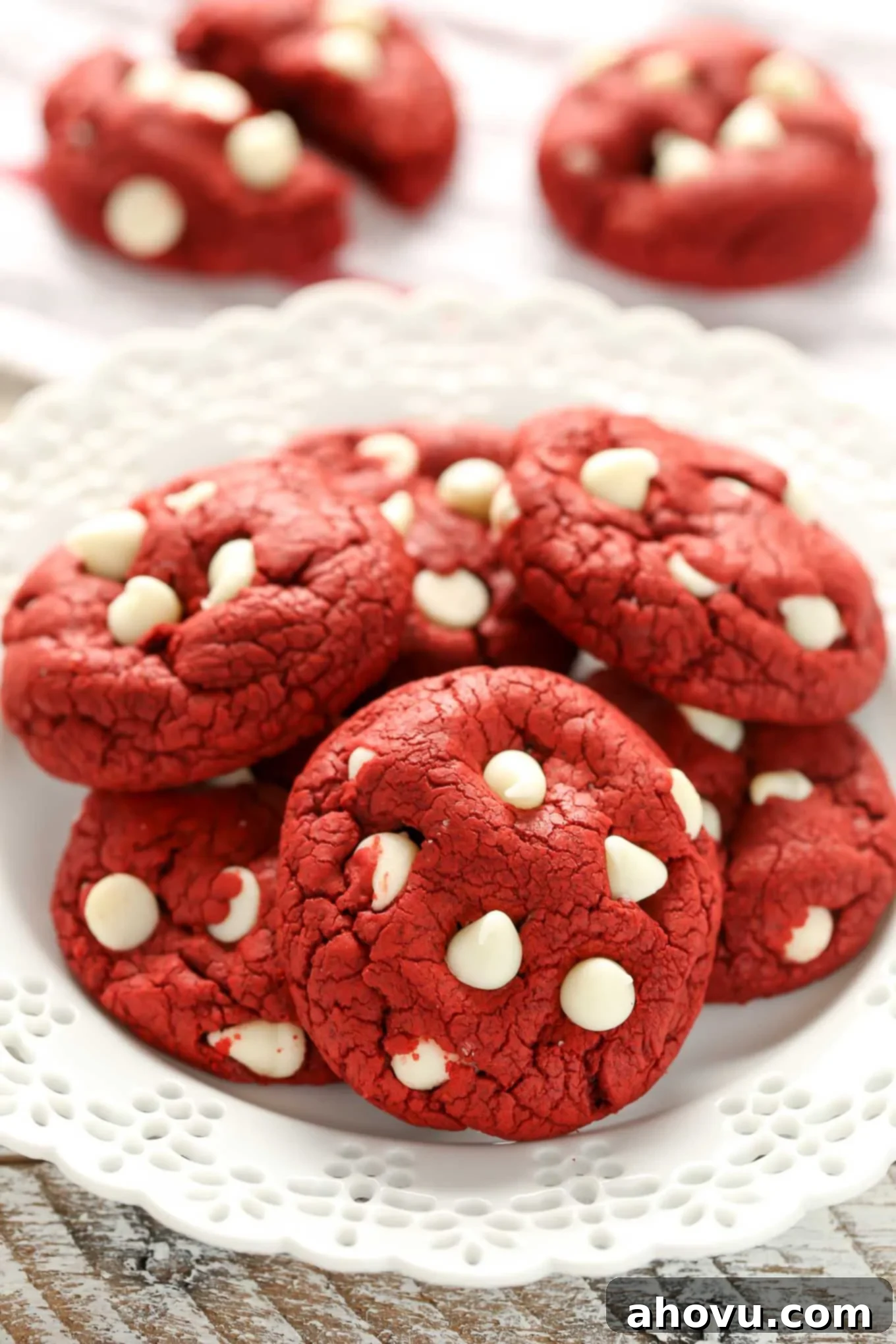 Effortless Red Velvet Delights 4 A white plate piled high with perfectly baked red velvet cookies, adorned with visible white chocolate chips. Two additional cookies are softly blurred in the background.