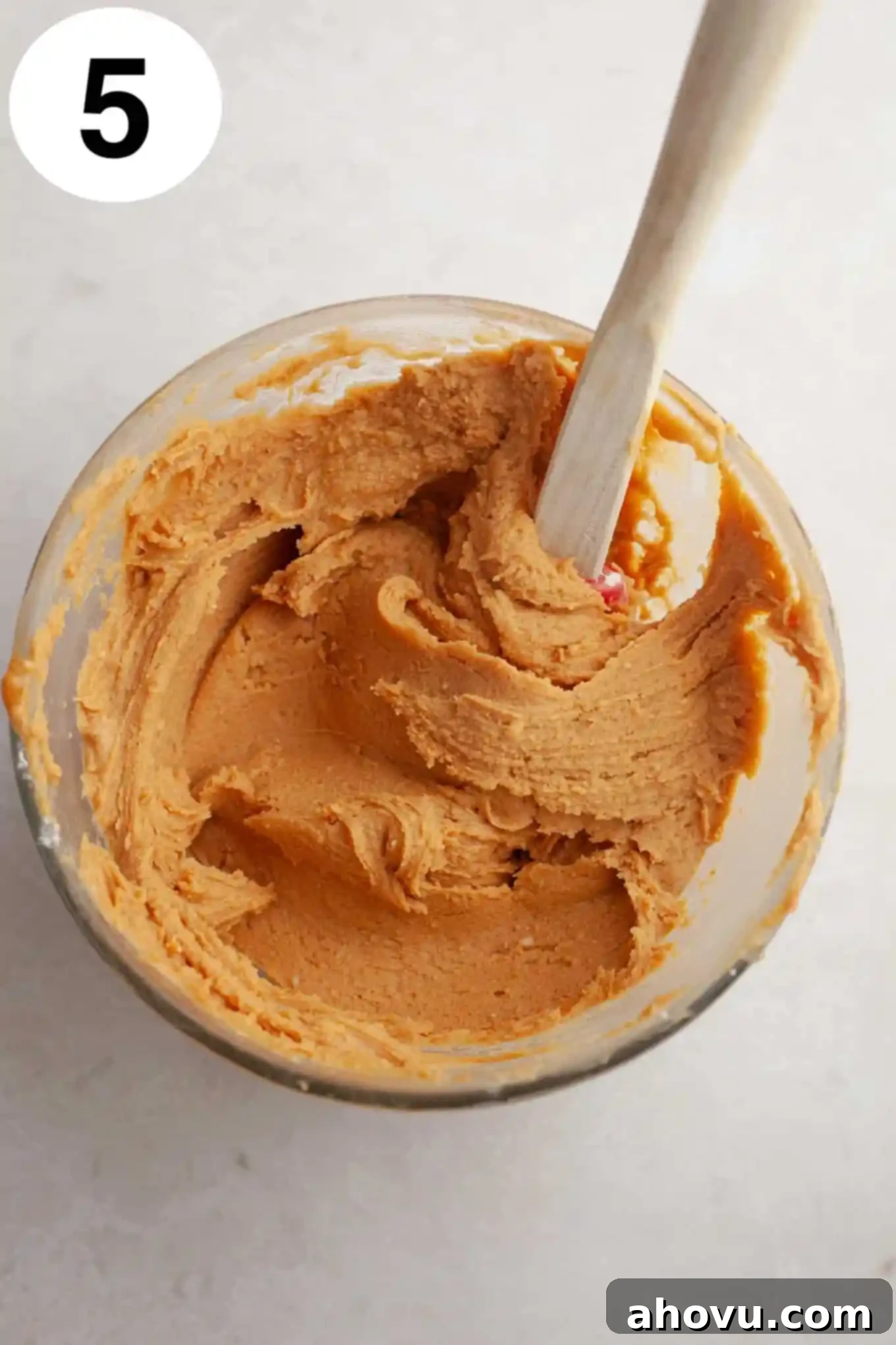 An overhead view of a smooth, rich peanut butter fudge mixture in a glass mixing bowl, with a spatula resting in it, ready for topping the cookies.