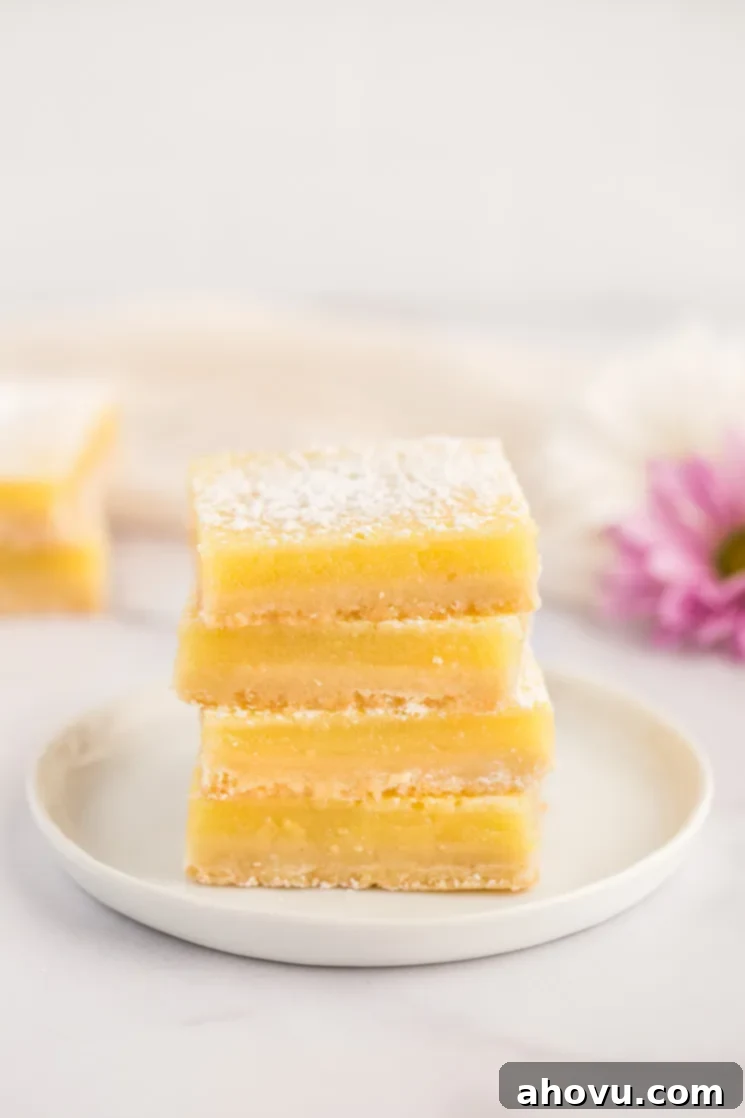 A stack of lemon bars on a white plate with more lemon bars and flowers in the background.