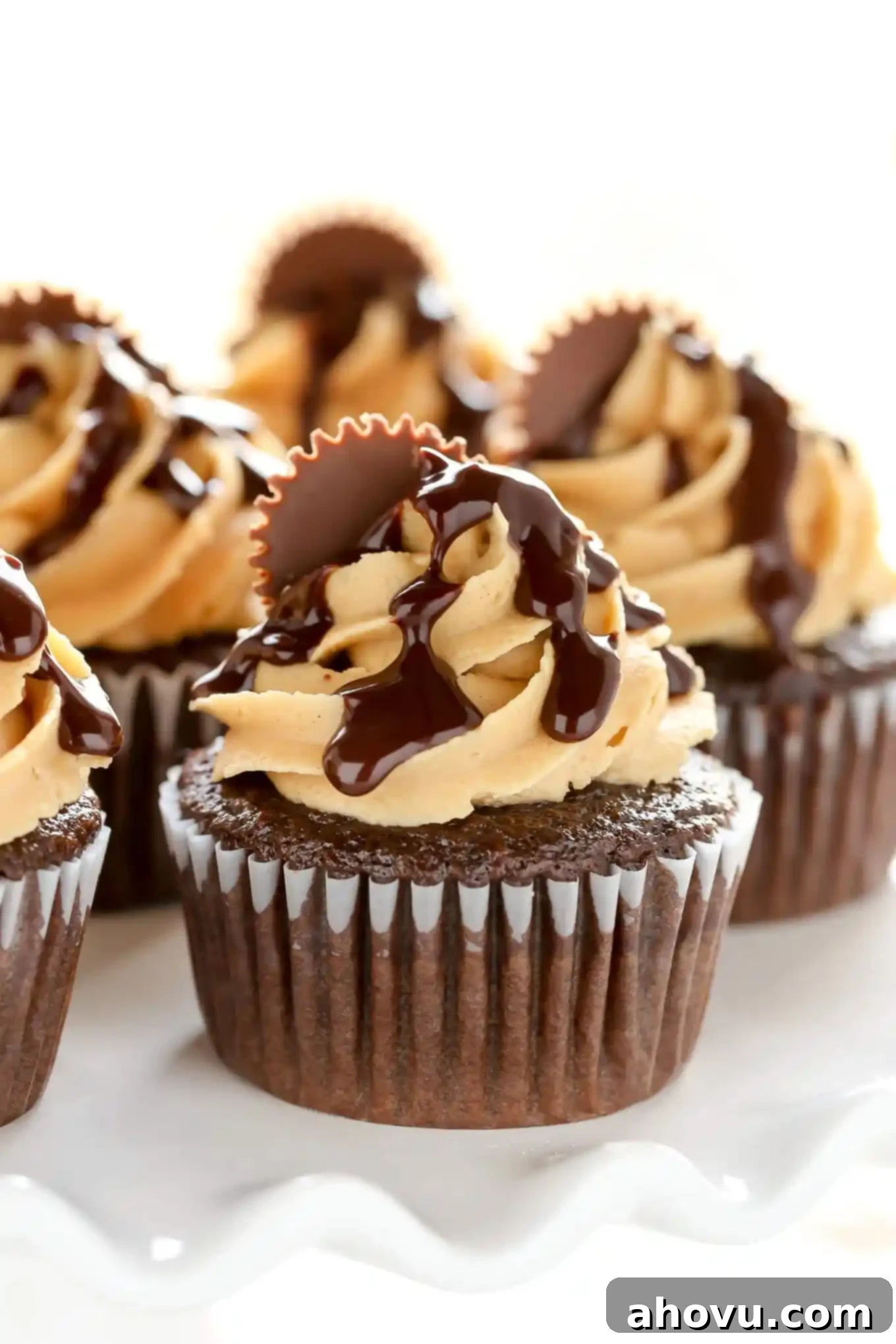 Close up of chocolate peanut butter cupcakes with rich ganache and peanut butter cup halves on a white cake stand, highlighting their irresistible texture and decadent toppings.