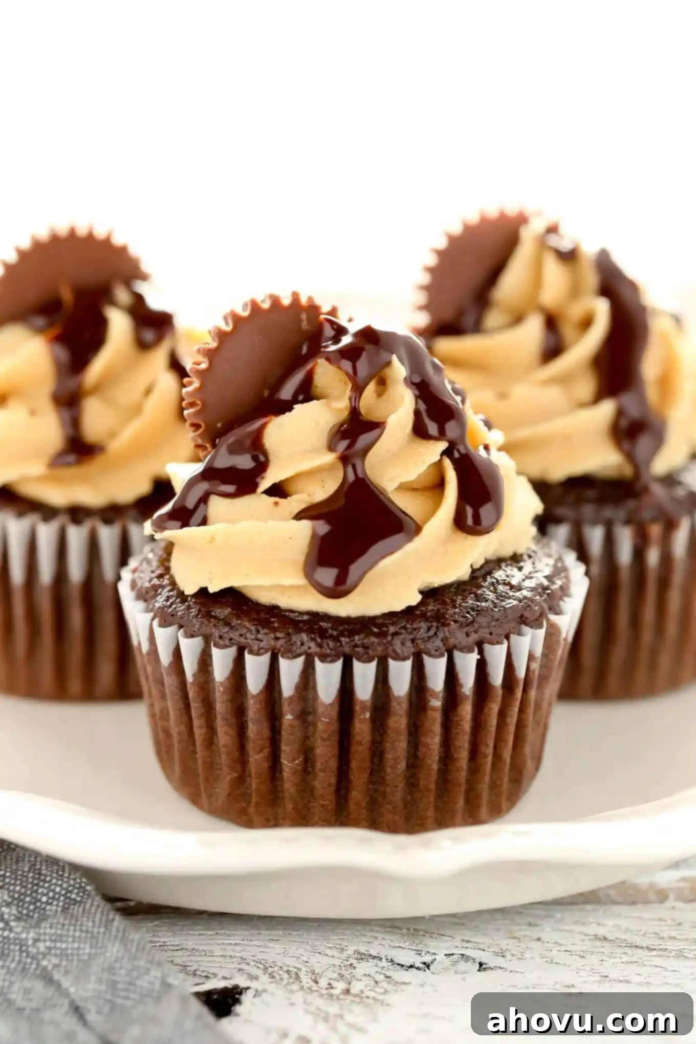 Close up shot of three chocolate peanut butter cupcakes, meticulously decorated with swirled frosting, chocolate ganache drips, and a halved Reese's Peanut Butter Cup, presented on a white plate for an enticing visual.