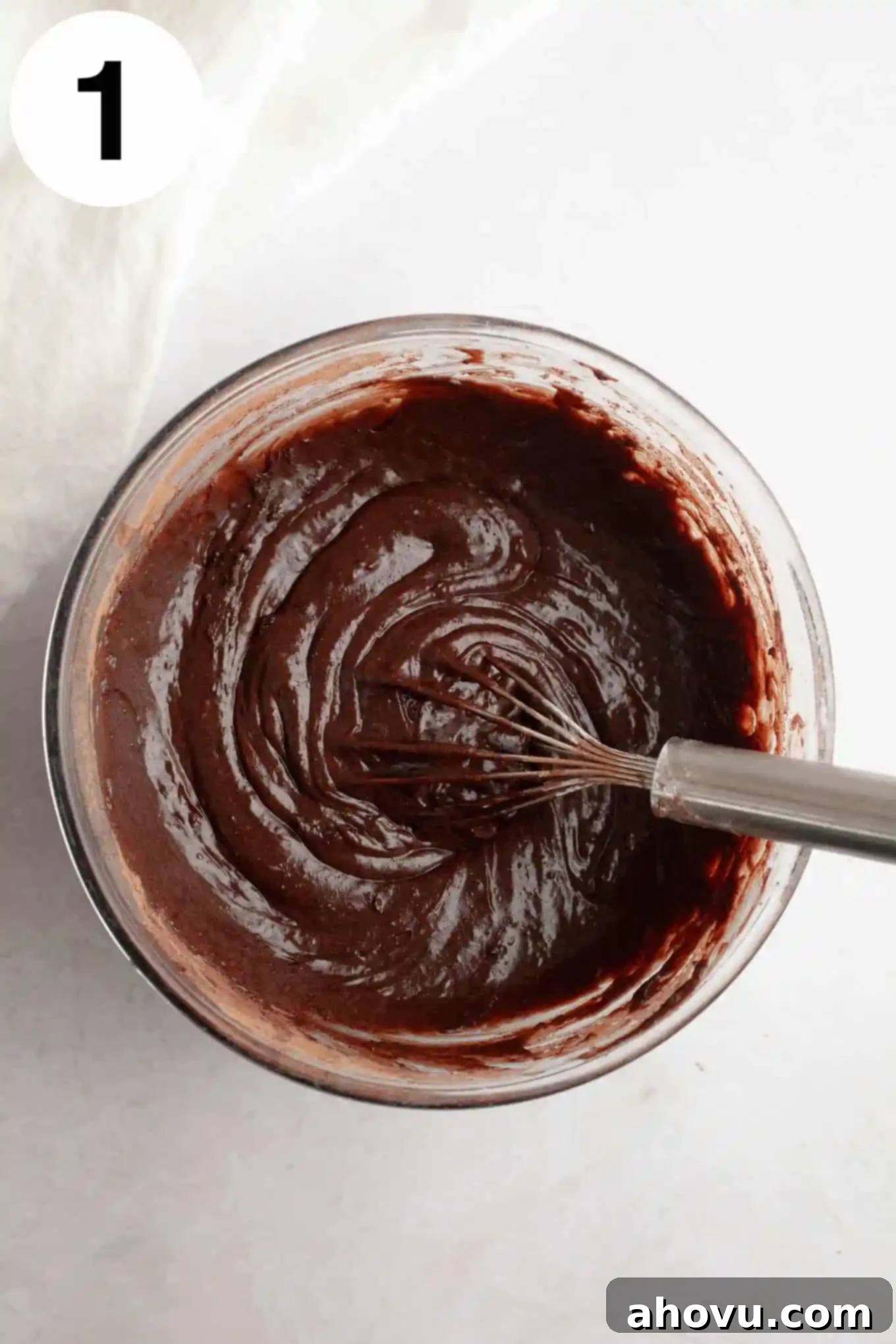 An overhead view of rich, dark brownie batter in a clear glass mixing bowl, being whisked to perfection.