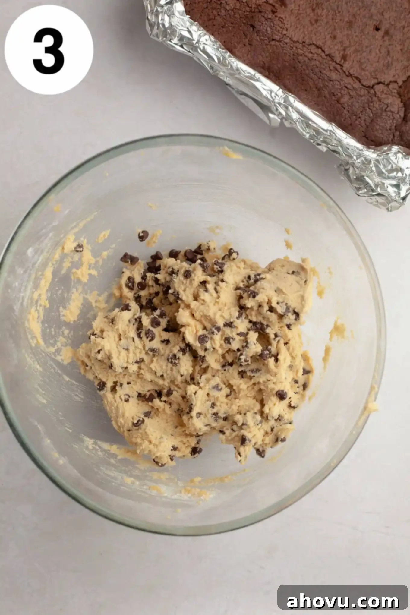 An overhead view of raw edible cookie dough in a clear glass mixing bowl, light brown and speckled with mini chocolate chips.