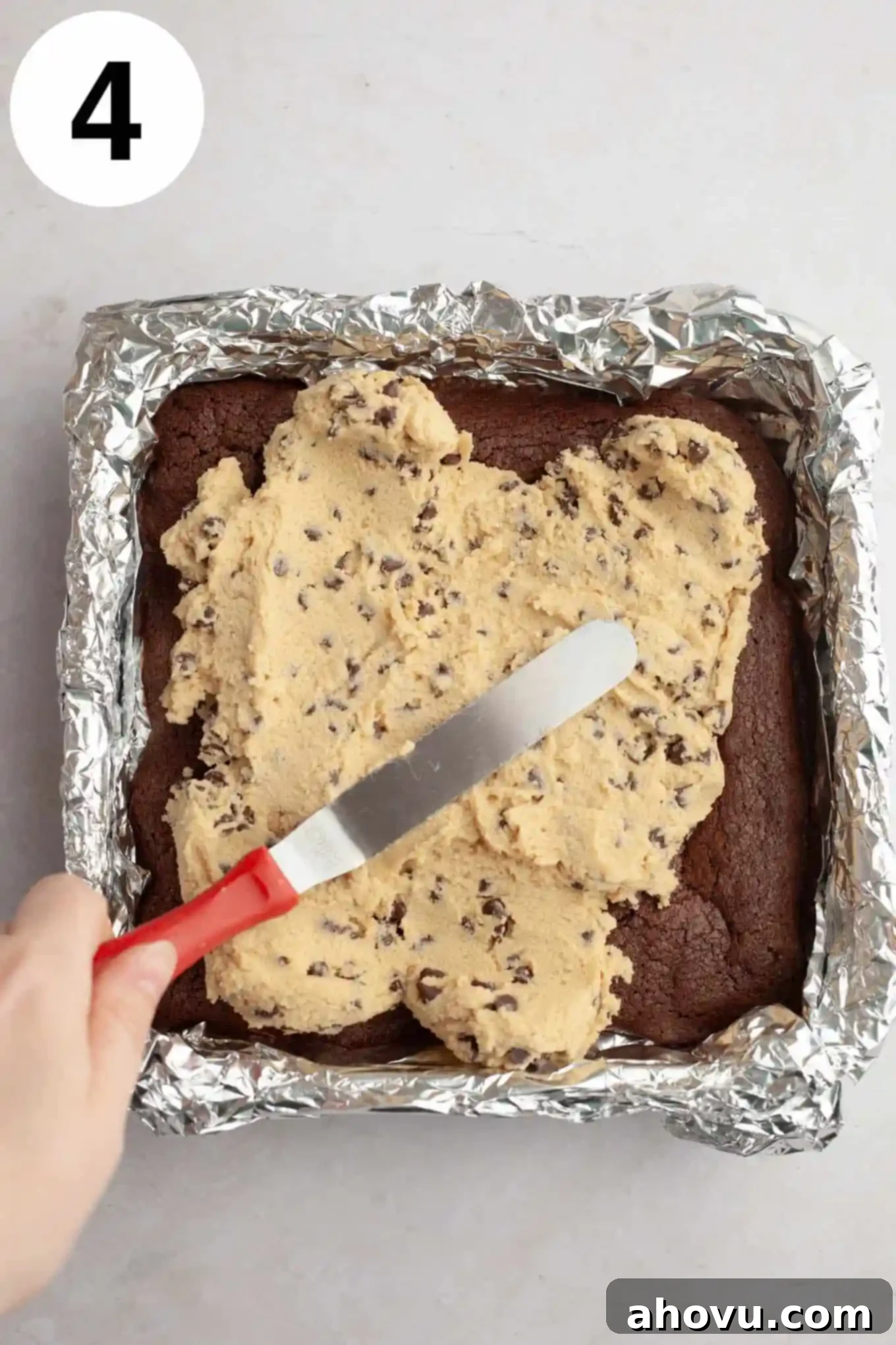 An overhead view of edible cookie dough being gently spread over the cooled brownie layer in a square pan.