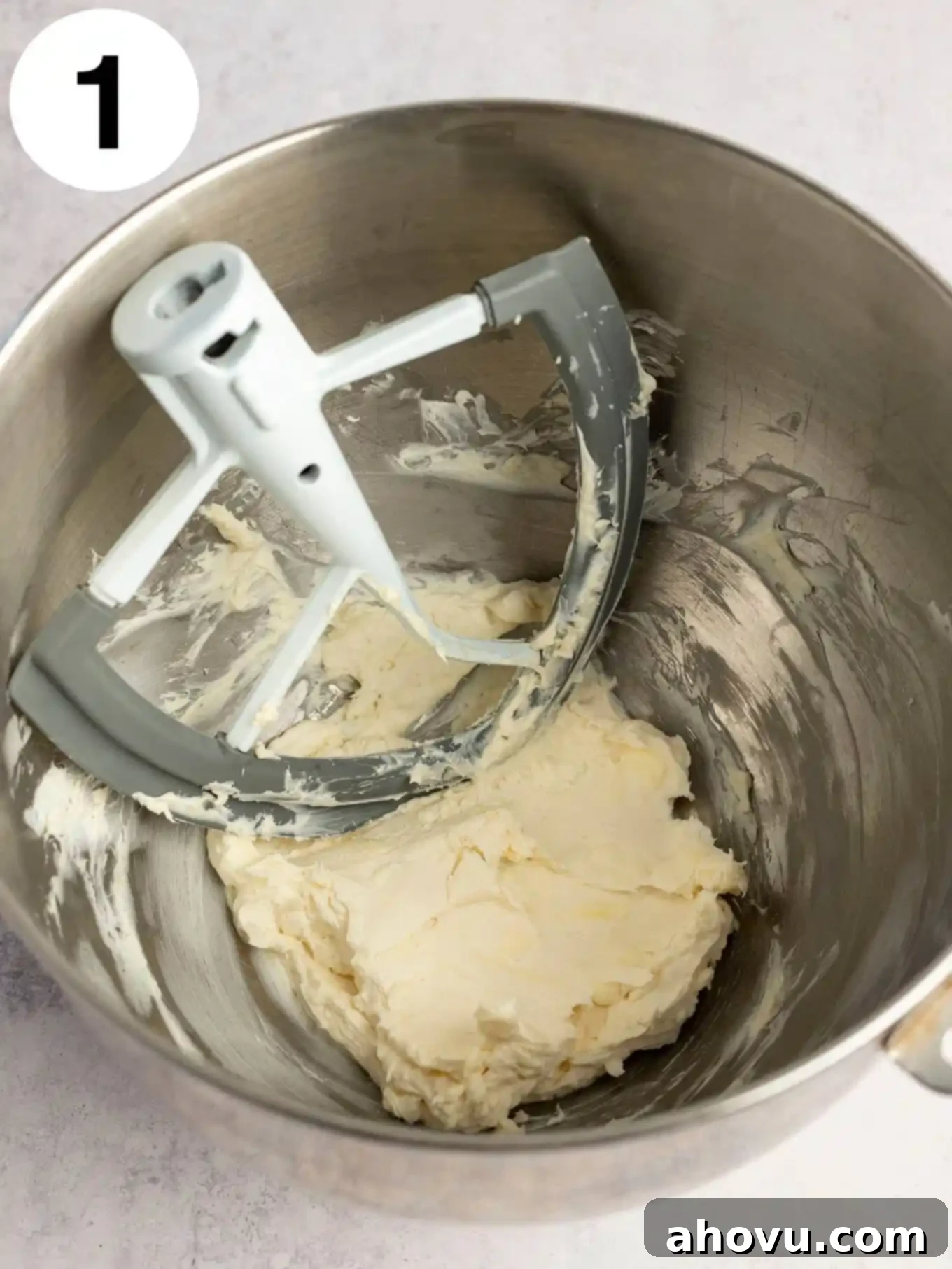 An overhead view of softened cream cheese and butter being creamed together in a mixing bowl with a paddle attachment.