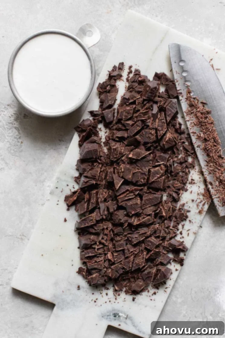 A marble cutting board displaying finely chopped semi-sweet chocolate next to a small cup filled with heavy whipping cream, ready for ganache preparation.