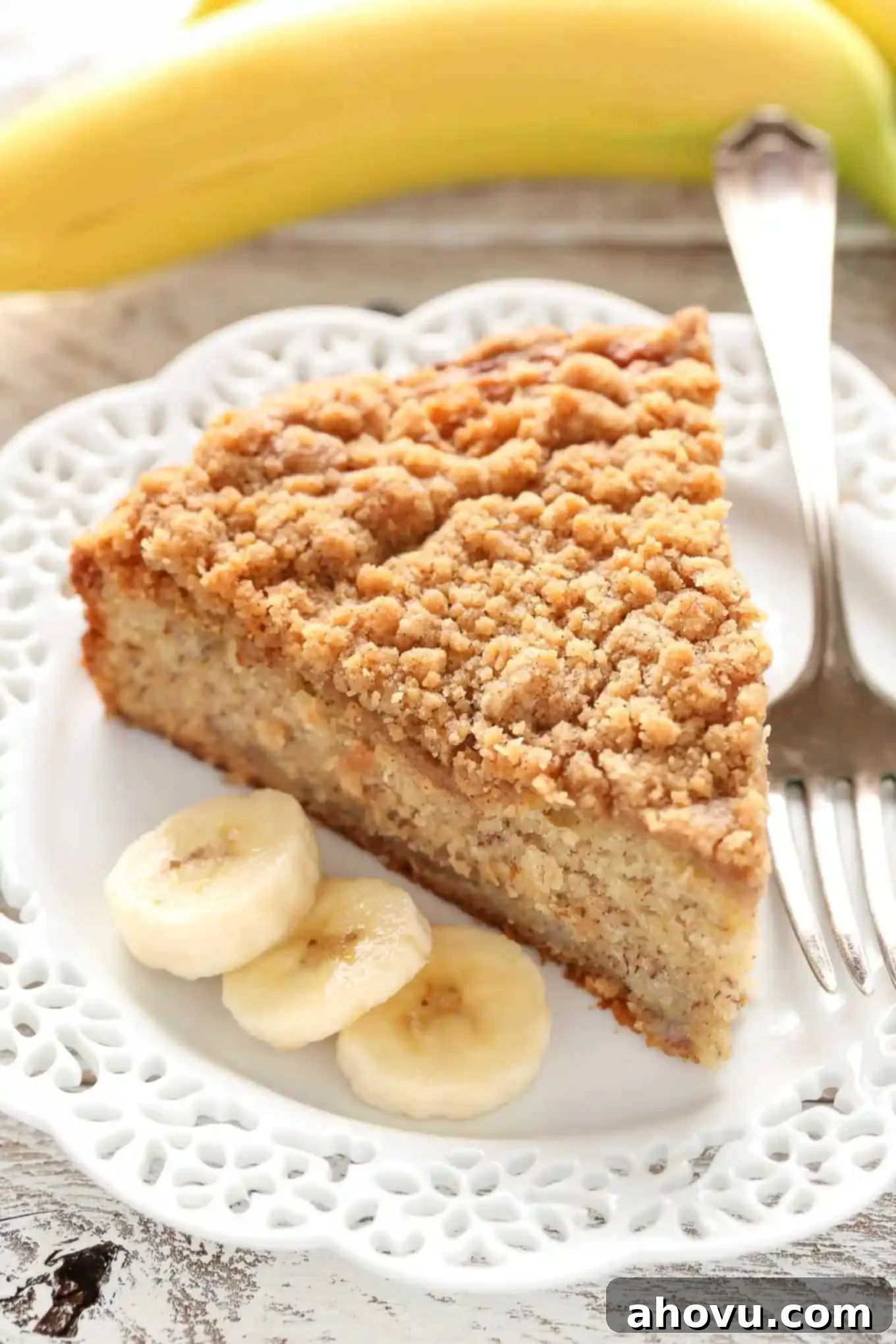 A slice of banana crumb cake, a fork, and three banana slices on a white plate. A banana rests in the background. 