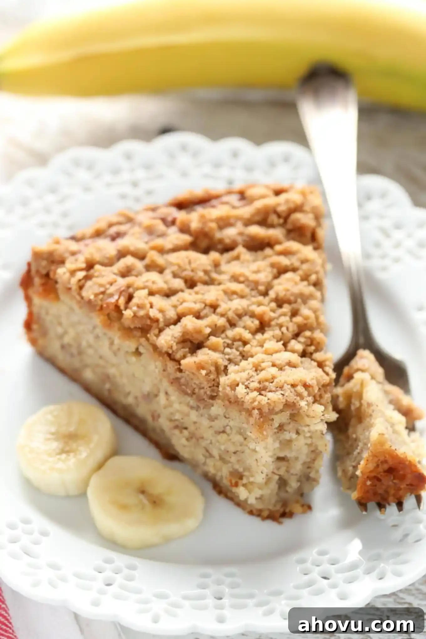 A slice of banana crumb cake, a fork, and three banana slices on a white plate. A banana rests in the background. 