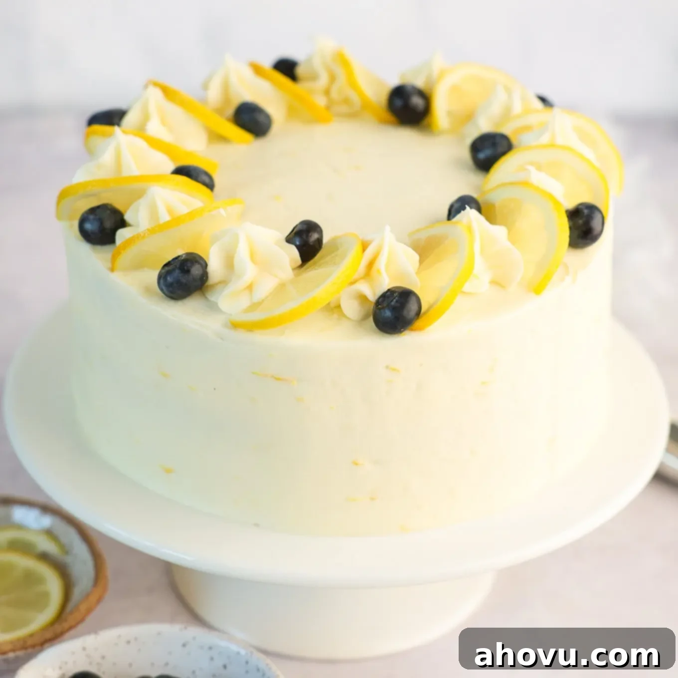 A beautifully decorated lemon blueberry cake, frosted and adorned with fresh lemon slices and vibrant blueberries on top, sitting on a cake stand.