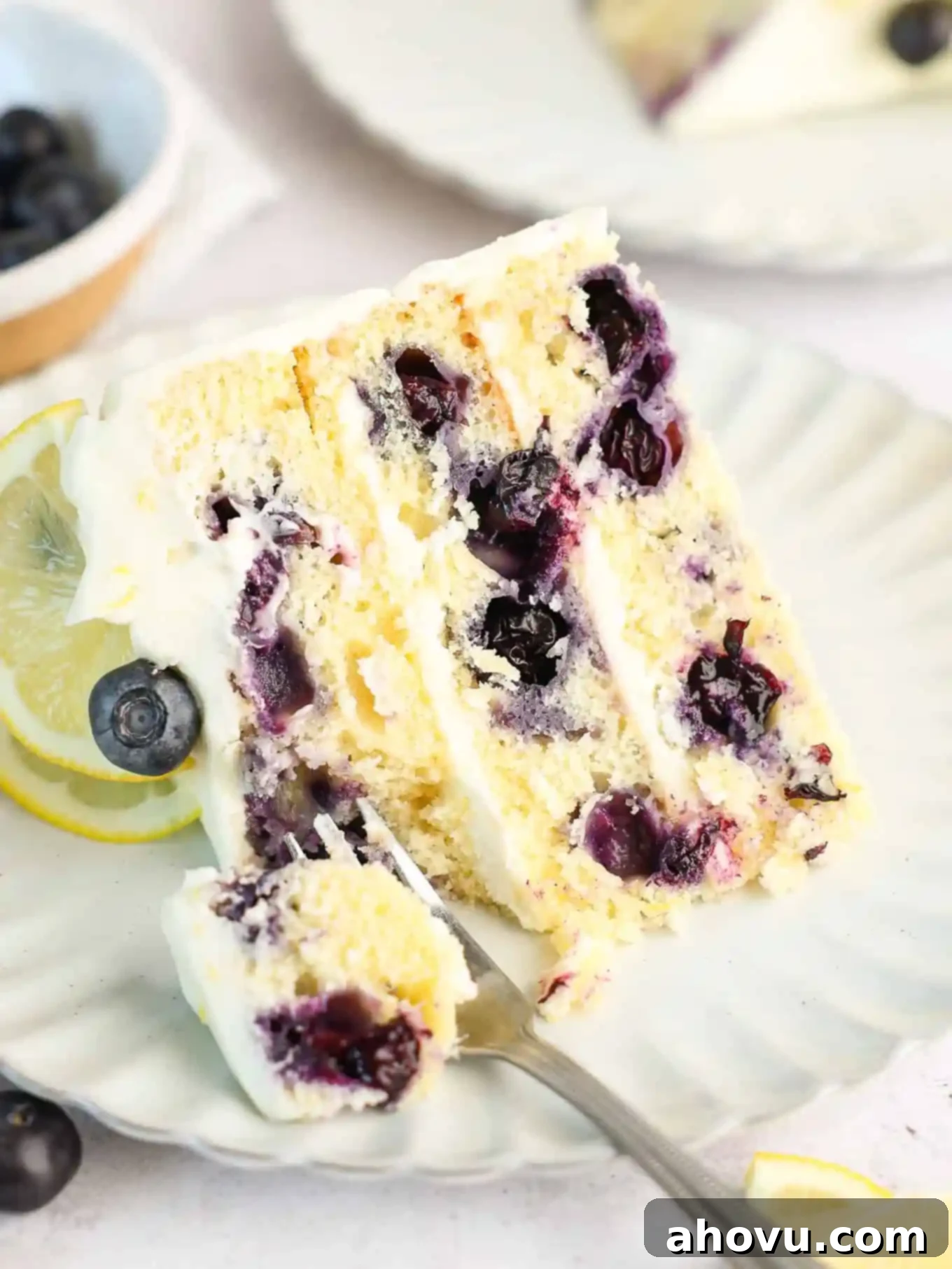 A single slice of moist lemon blueberry cake, generously frosted, presented on a white dessert plate with a fork.