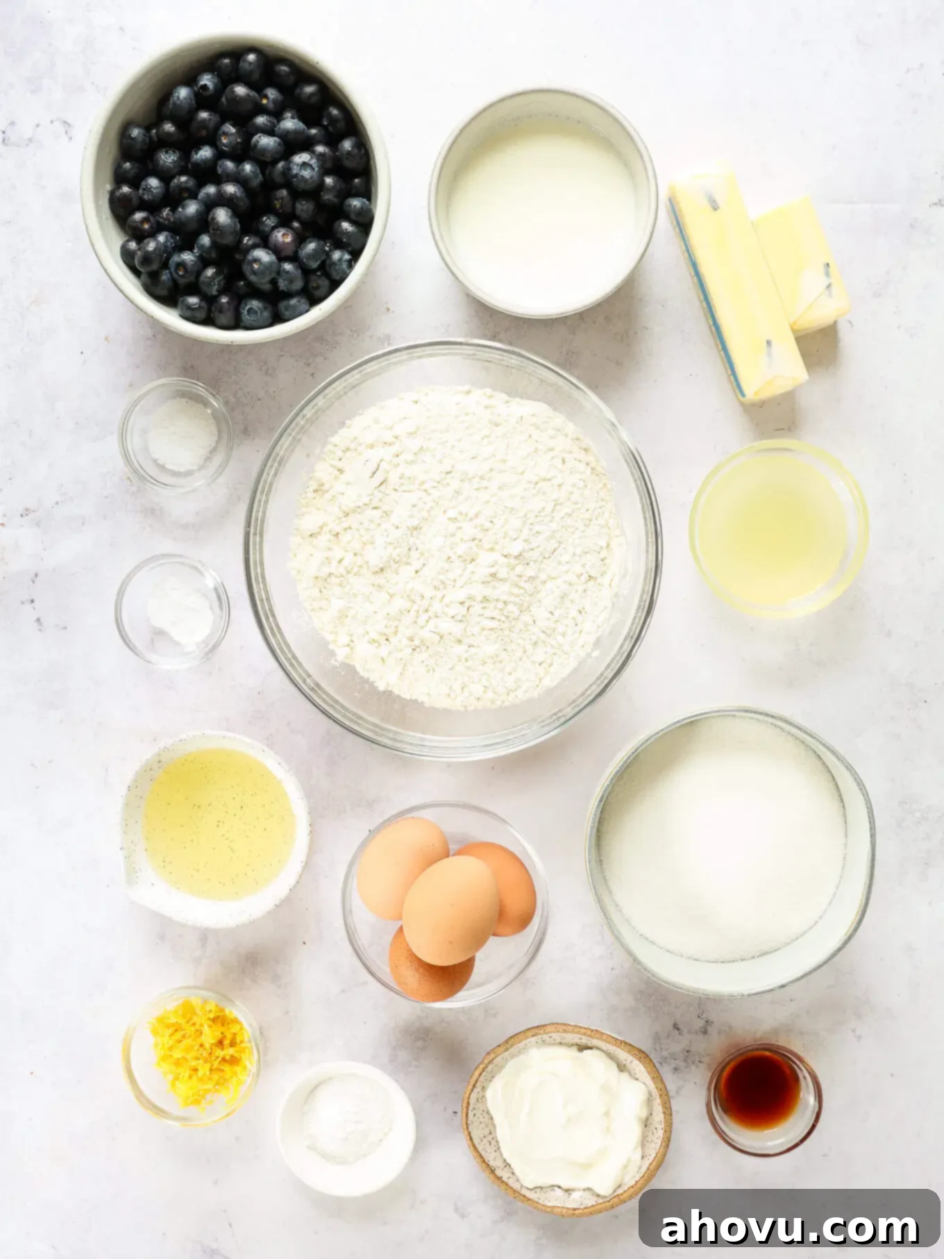 A collection of fresh ingredients laid out on a marble counter, including lemons, blueberries, flour, butter, and other baking essentials for lemon blueberry cake.