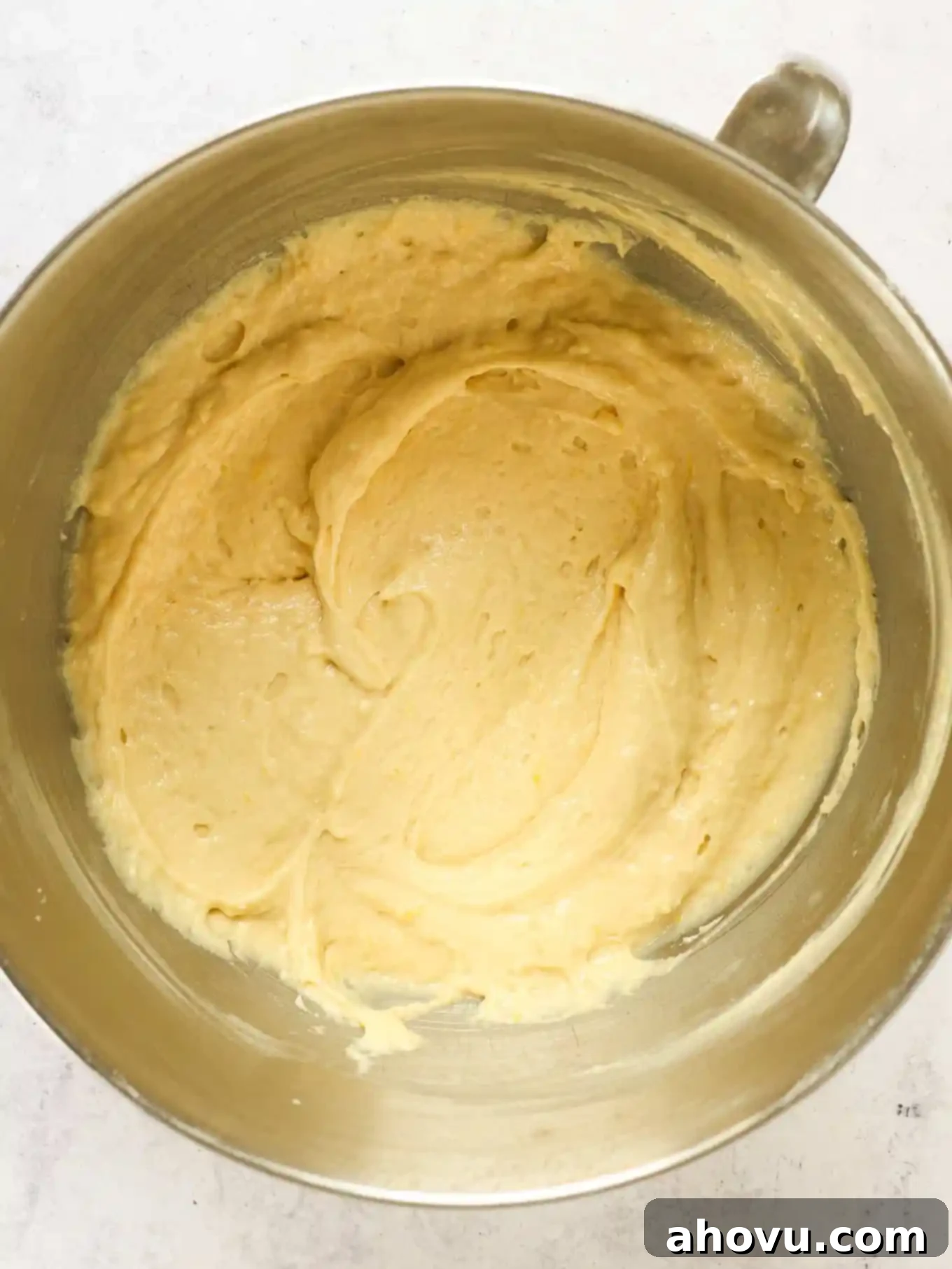 An overhead shot of glossy, pale yellow lemon cake batter in a stainless steel mixing bowl, ready for the blueberries.
