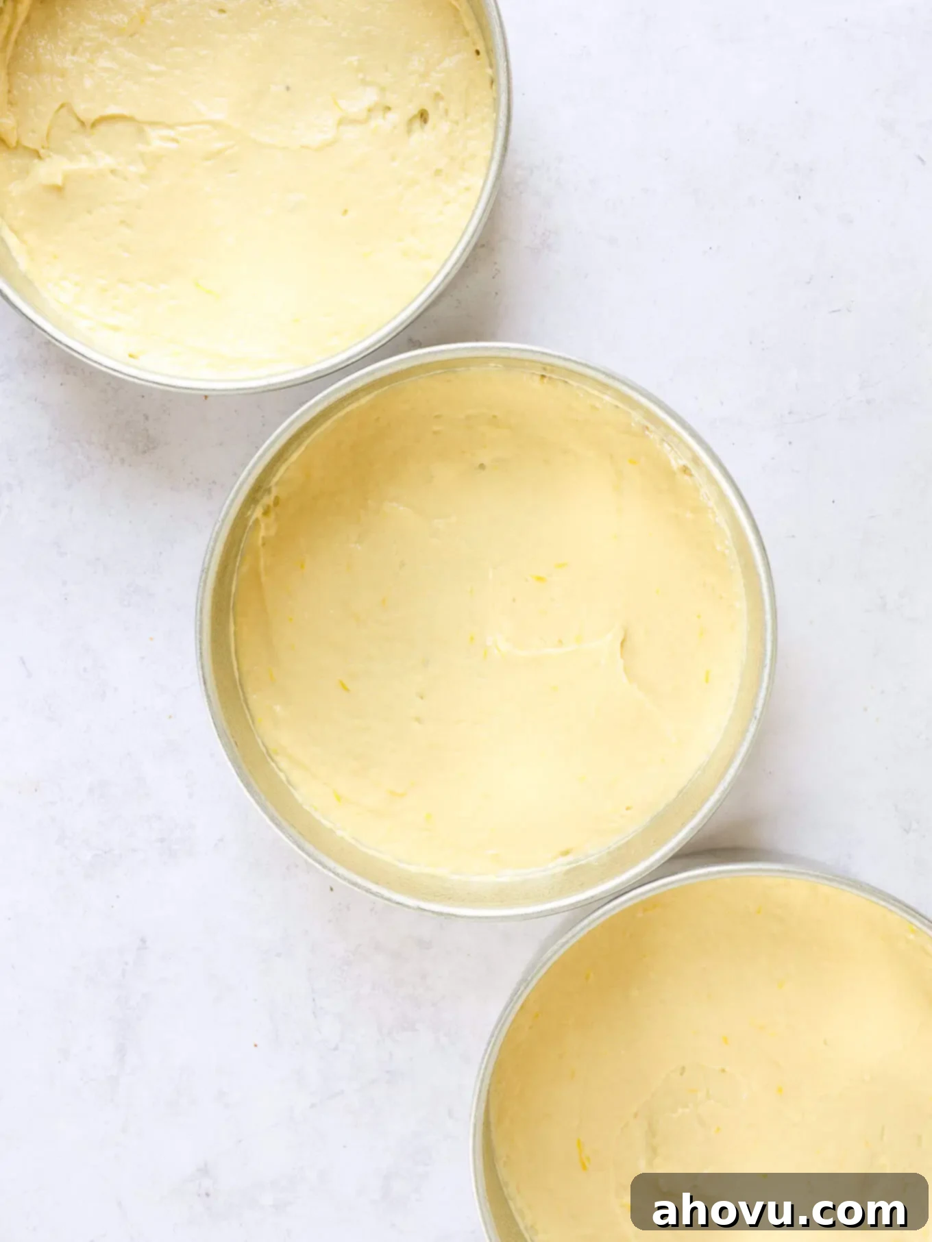 Three empty cake pans with a small, thin layer of plain lemon cake batter spread at the bottom of each.