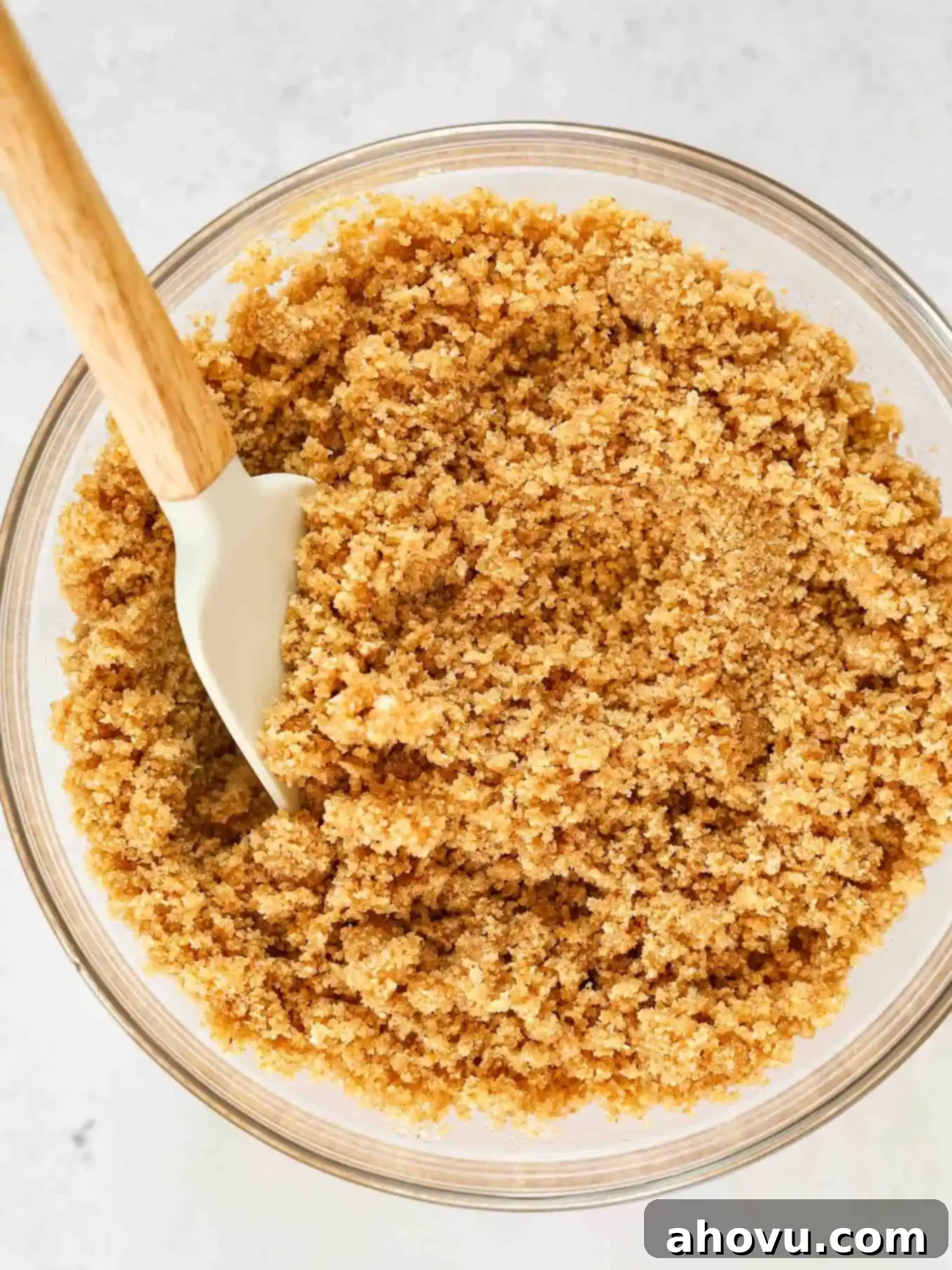 An overhead view of the graham cracker crust mixture, a golden brown blend of crumbs, sugar, and melted butter, in a glass mixing bowl.