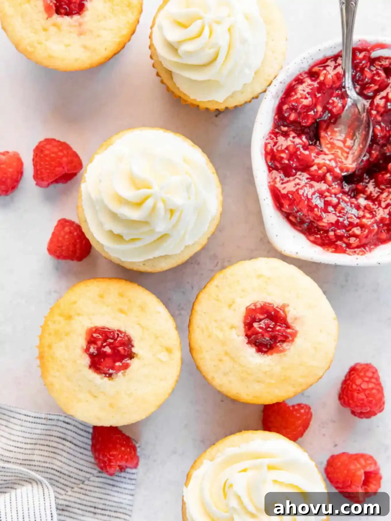 An overhead shot of several perfectly filled white cupcakes, with a bowl of raspberry filling nearby, inviting a taste.