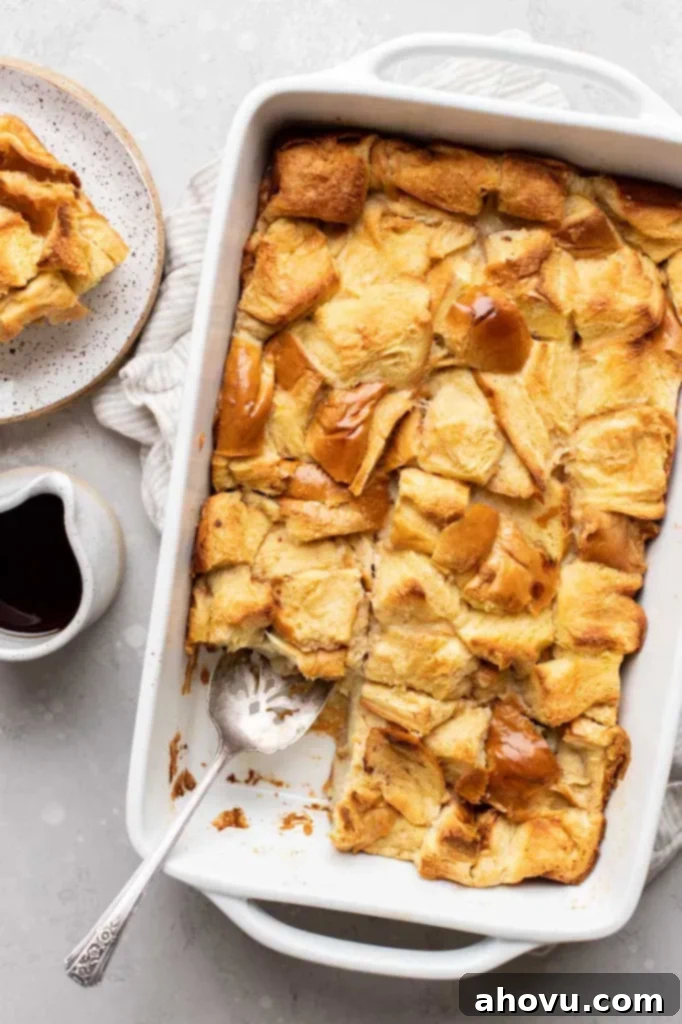 An overhead view of golden-brown baked bread pudding in a 9x13 pan, with a slice removed and a small container of syrup beside it, ready for serving.