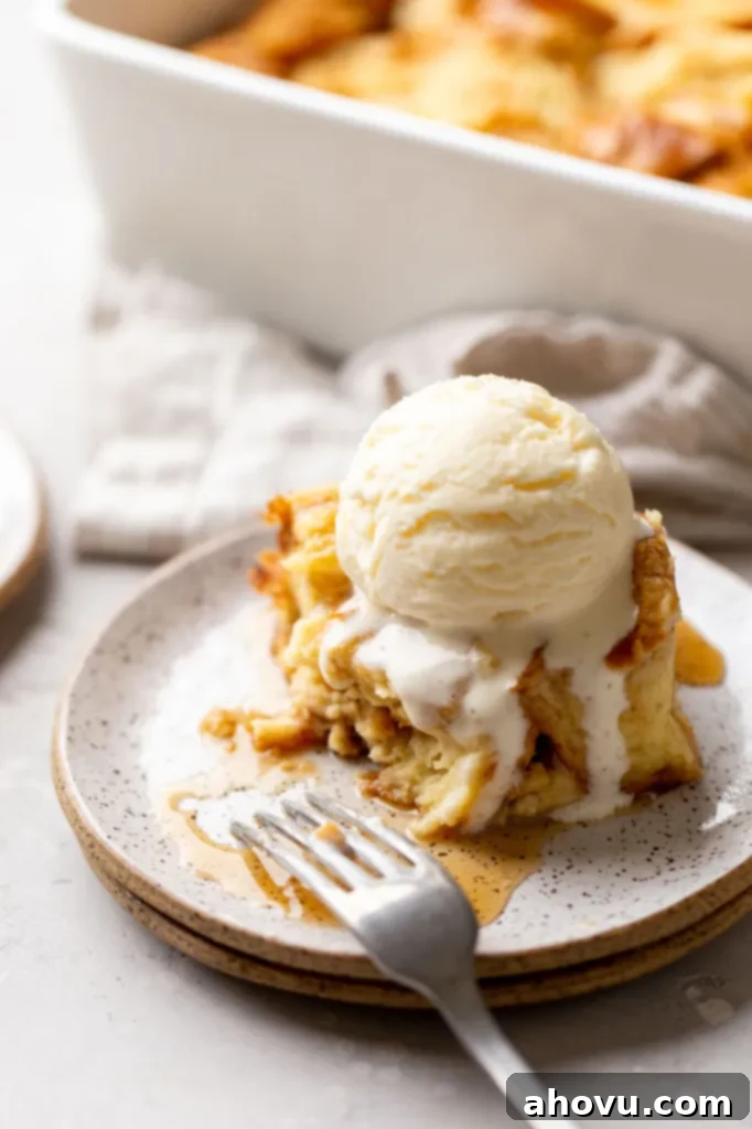 A close-up of a slice of bread pudding topped with melting vanilla ice cream, with a bite taken to reveal the soft, custardy interior and golden-brown crust.