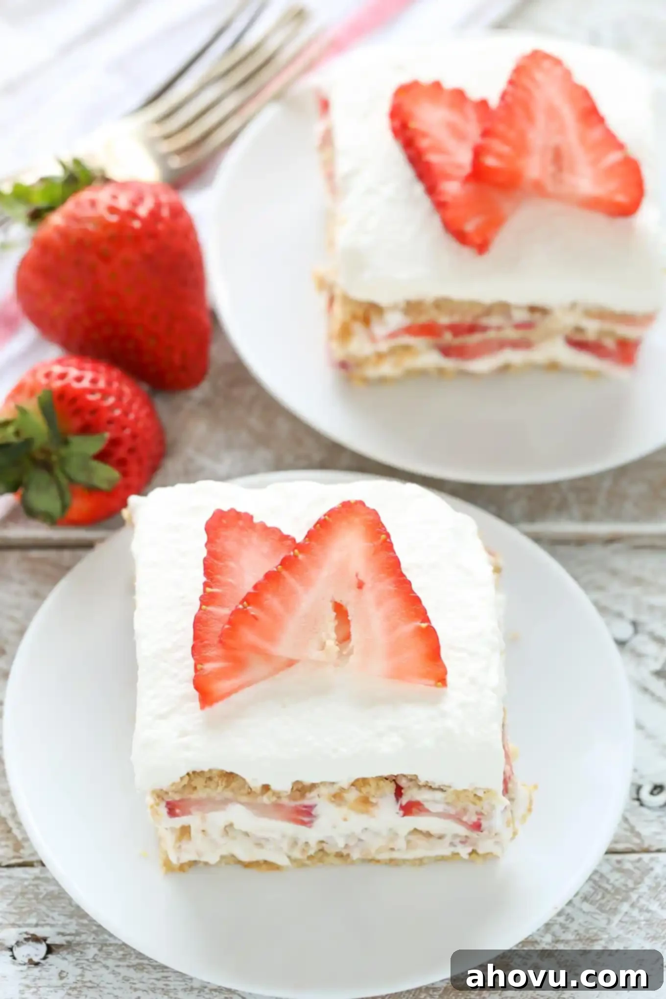 Two slices of strawberry icebox cake on white dessert plates. Two fresh berries and a fork rest on the side. 